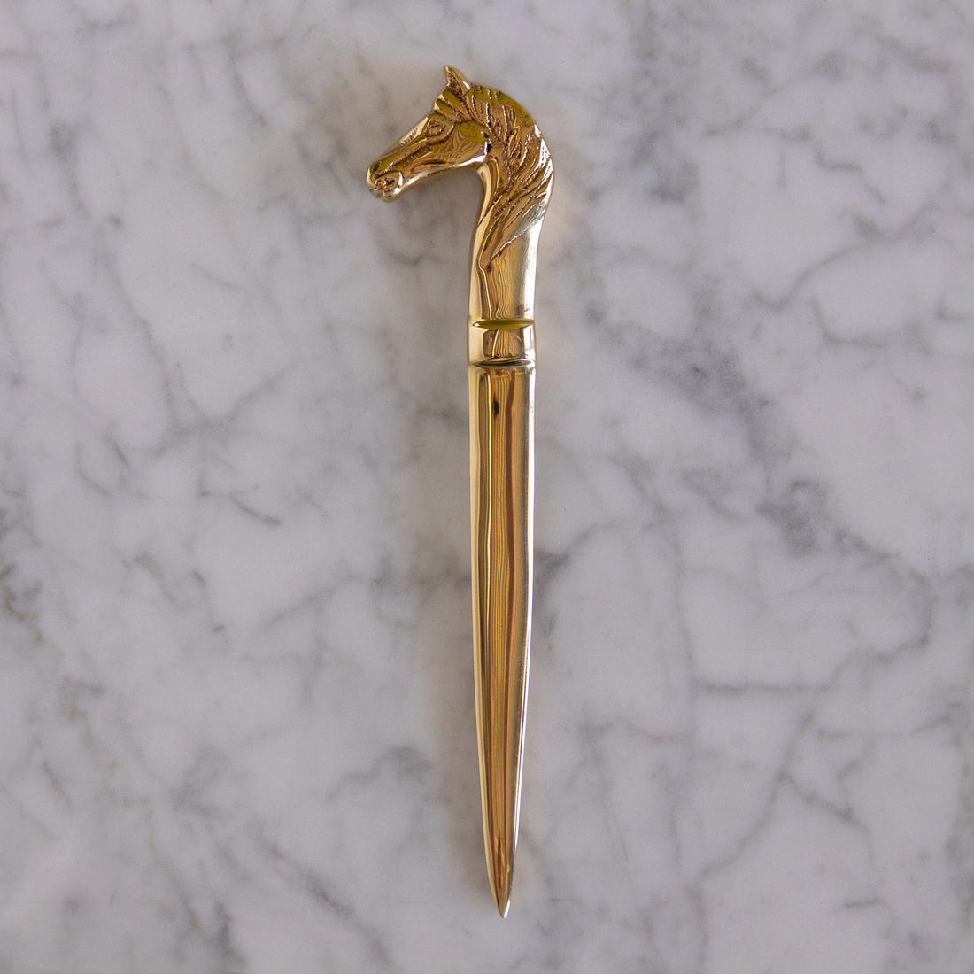 A polished gold-toned metal letter opener with a sculpted horse head handle, displayed vertically on a white and gray veined marble surface.