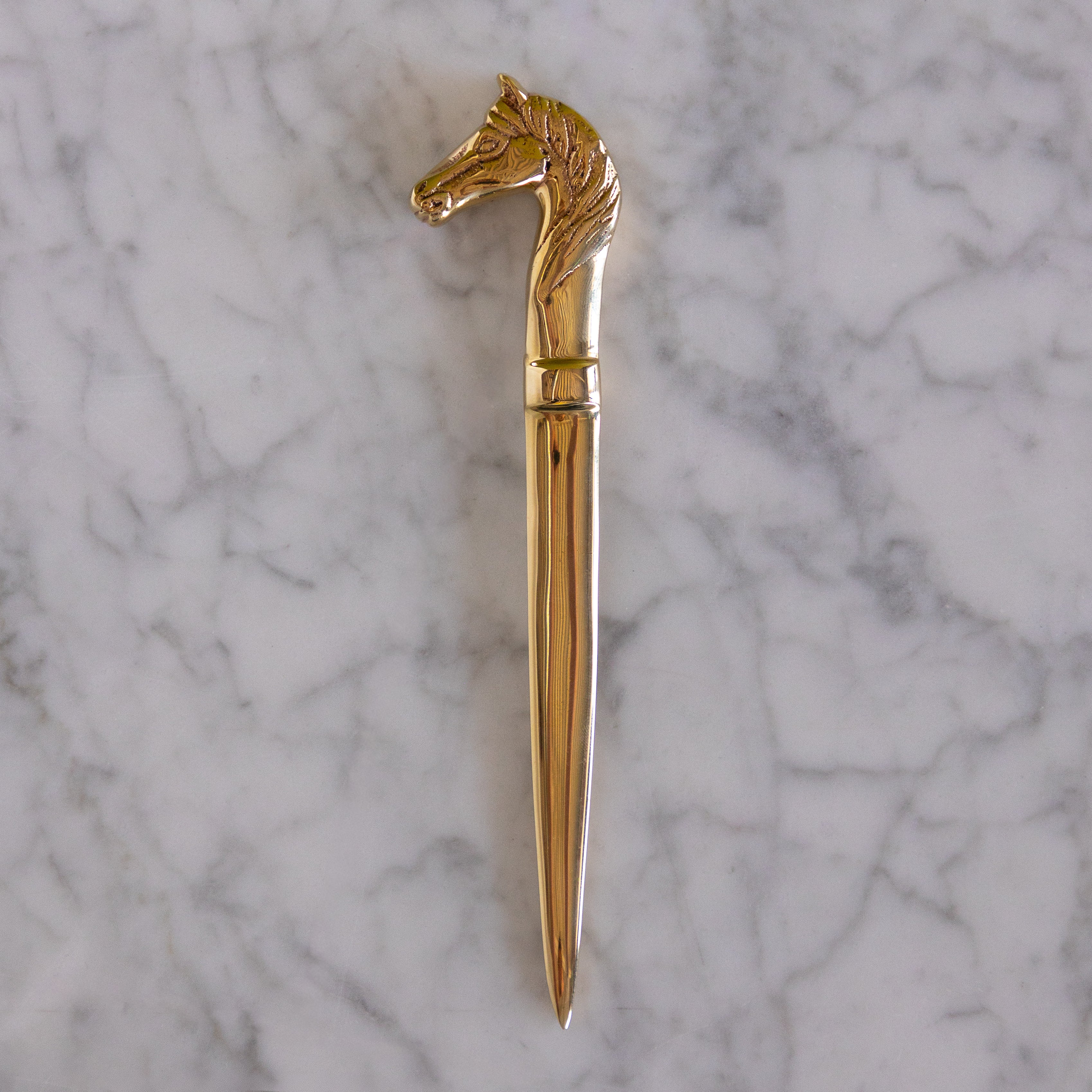 A polished gold-toned metal letter opener with a sculpted horse head handle, displayed vertically on a white and gray veined marble surface.