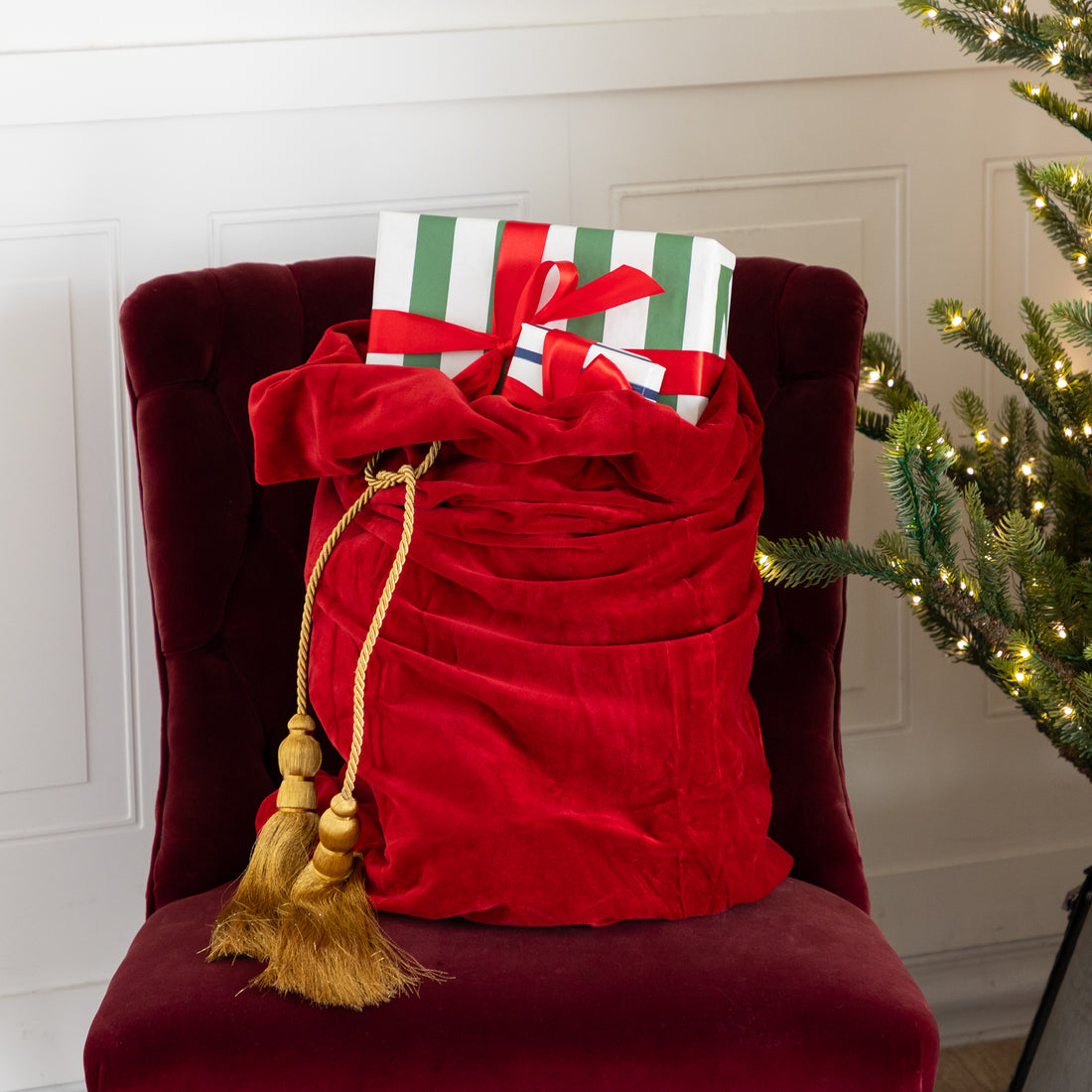 Red Velvet Santa Bag with two gold tassels and gifts inside, on a red chair next to a decorated Christmas tree.