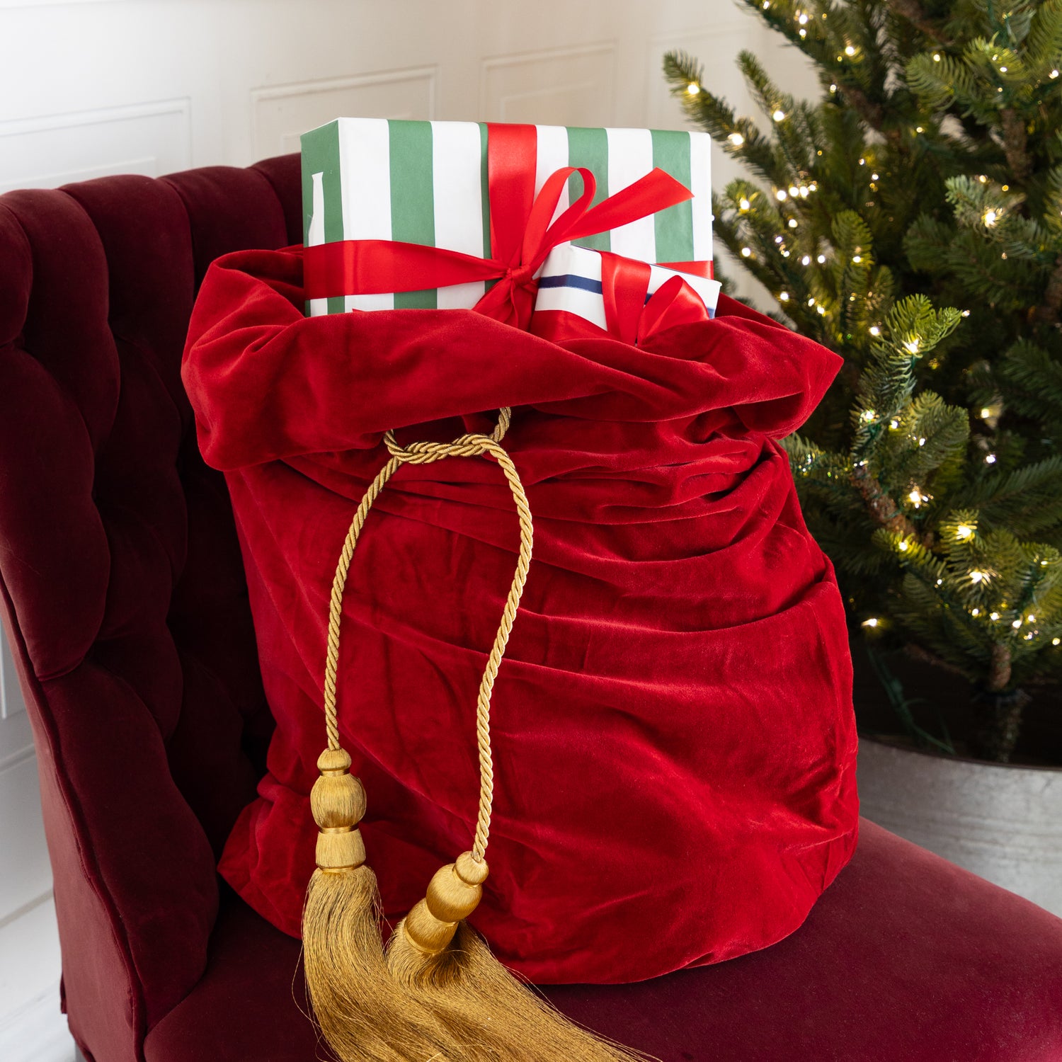 Red Velvet Santa Bag with two gold tassels and gifts inside, on a red chair next to a decorated Christmas tree.