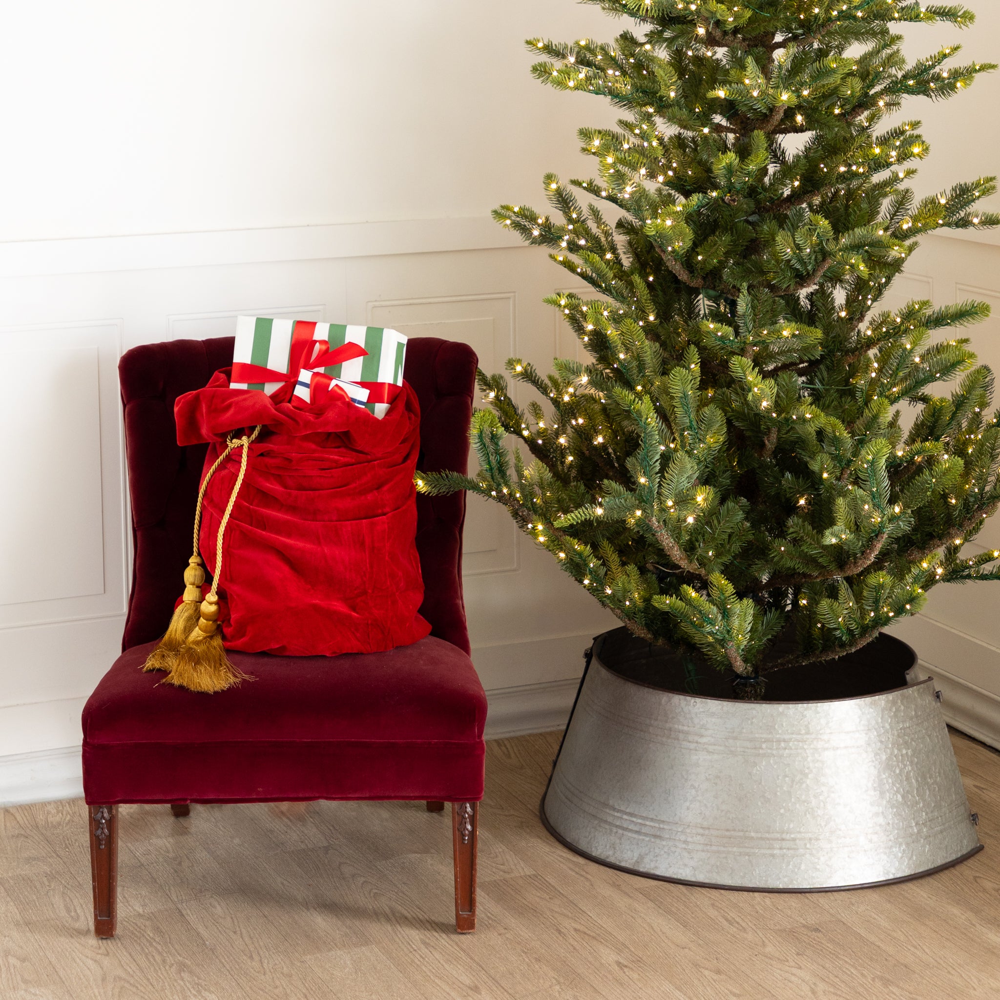 Red Velvet Santa Bag with two gold tassels and gifts inside, on a red chair next to a decorated Christmas tree.