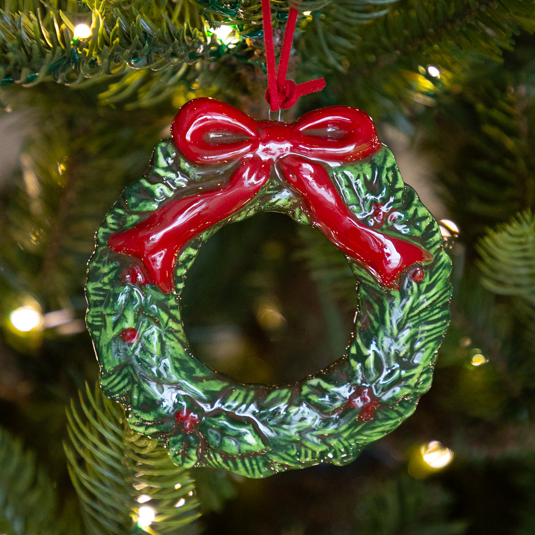 A green holly wreath Christmas ornament with a glossy red bow and red hanging string, shown hanging on a Christmas tree with warm lights in the background.