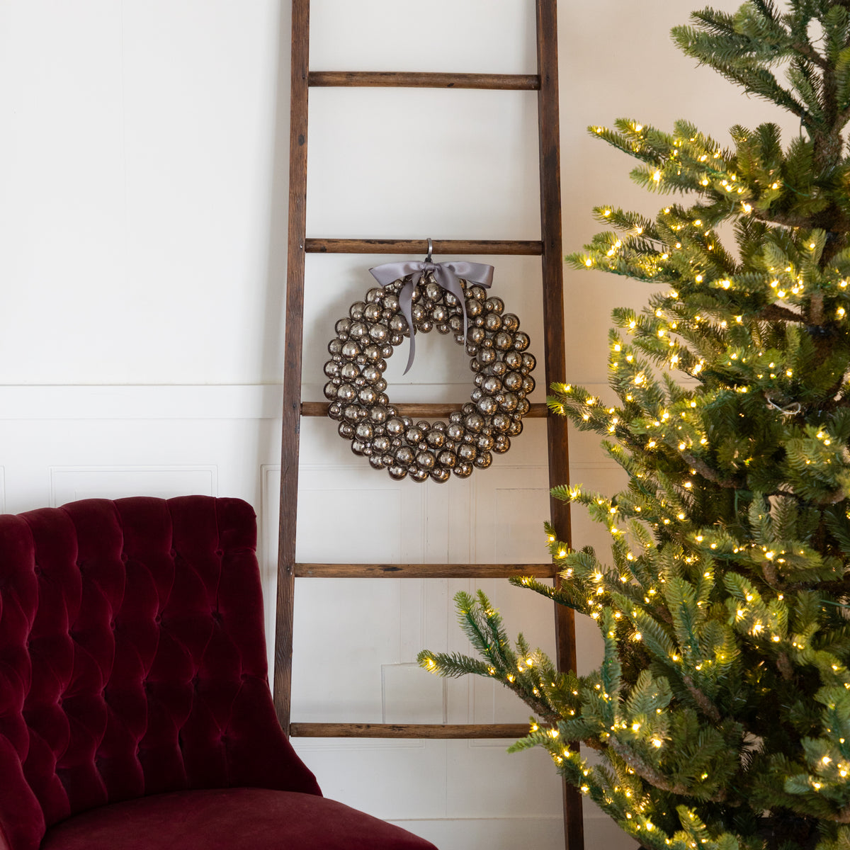  Antique Silver Bubble Wreath with a silver bow hanging on a wooden ladder next to a Christmas tree with lights and a red velvet chair, against a white wall.