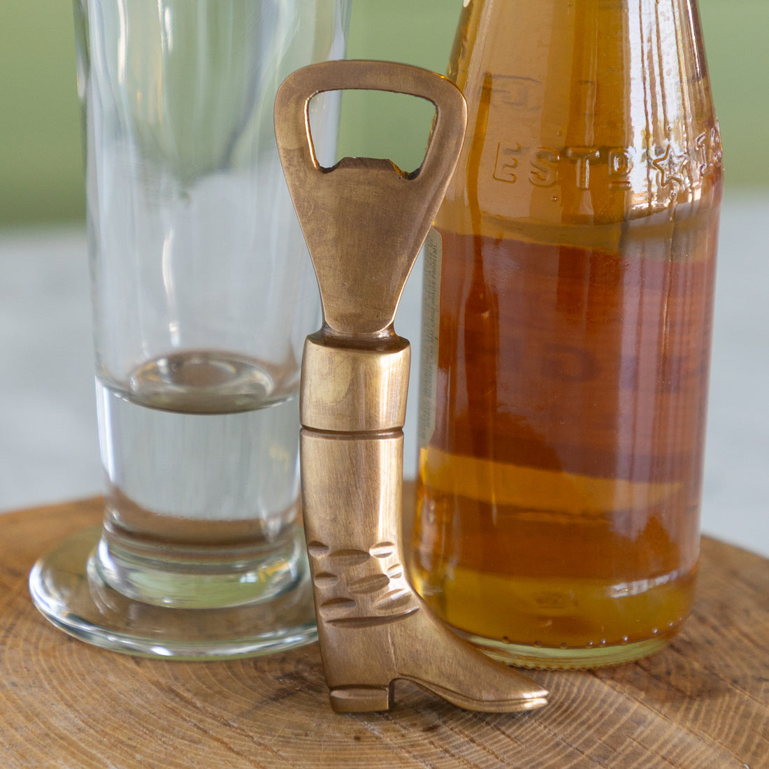 A brass bottle opener shaped like a boot, standing upright on a wooden surface between a clear glass and a brown glass bottle.