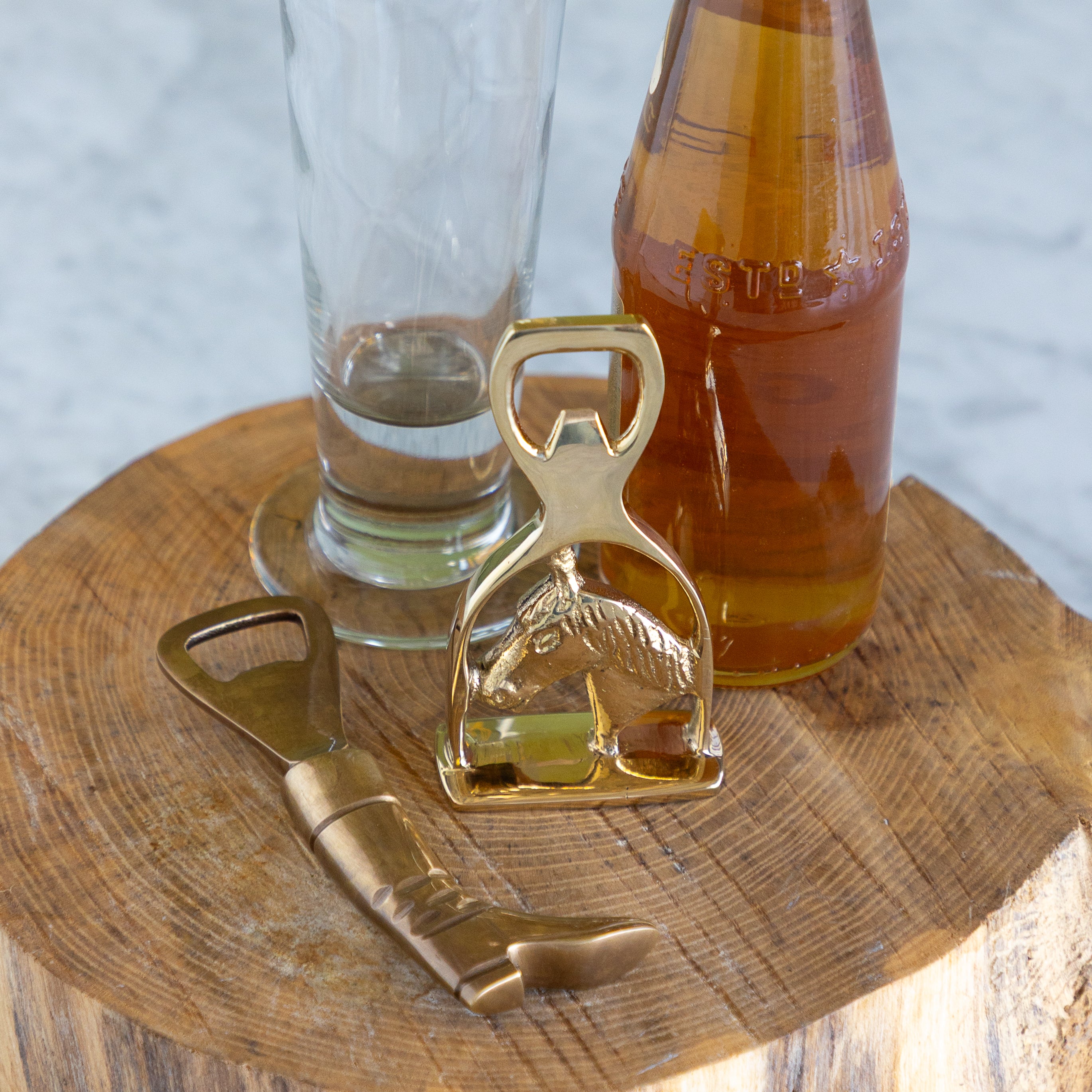 A brass boot shaped bottle opener next to a brass horse in a stirrup shaped bottle opener on a piece of wood next to a bottle and a glass. 