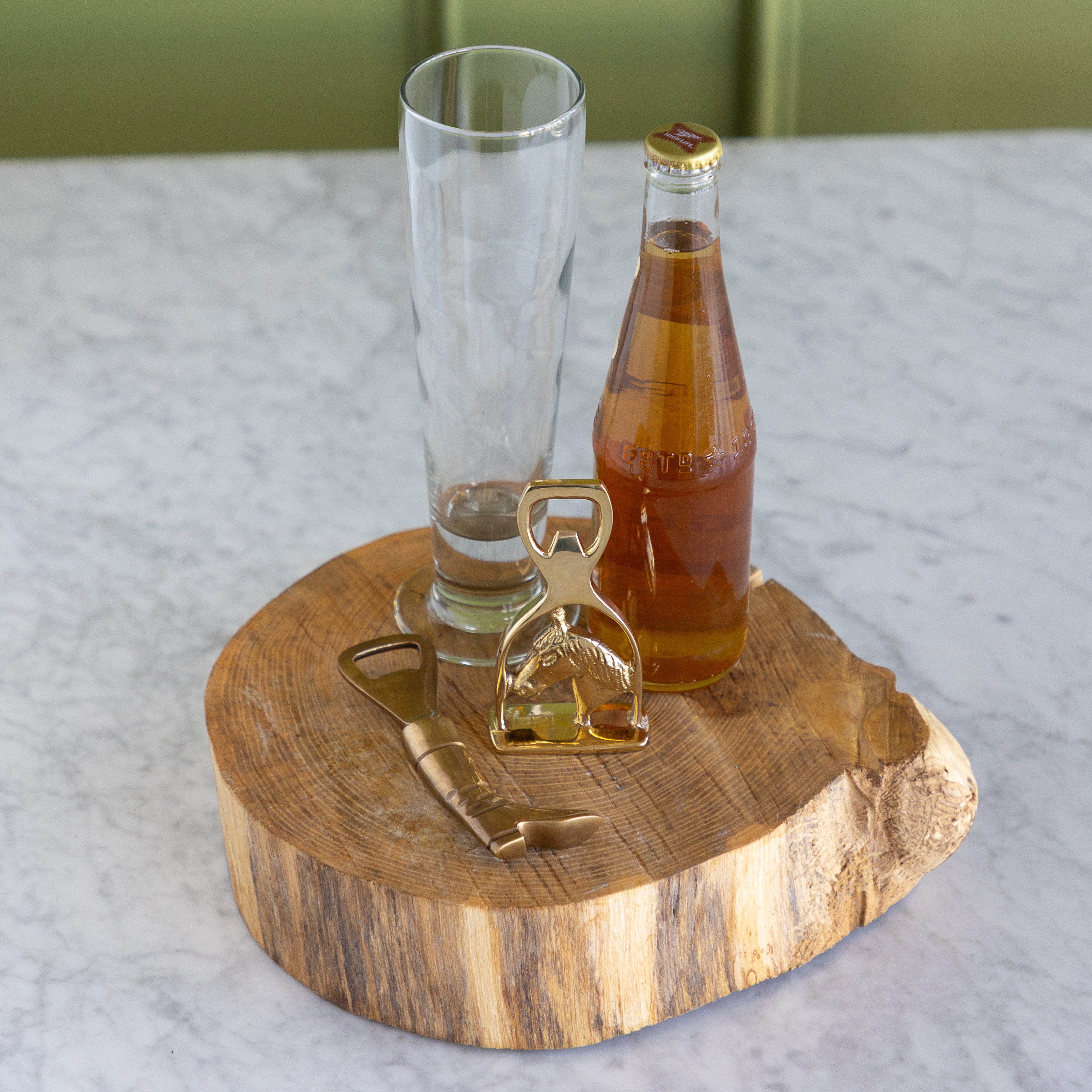 A rustic round wooden slice serving tray on a white marble countertop, styled with a clear textured drinking glass, a glass bottle of amber liquid, and two brass bottle openers.