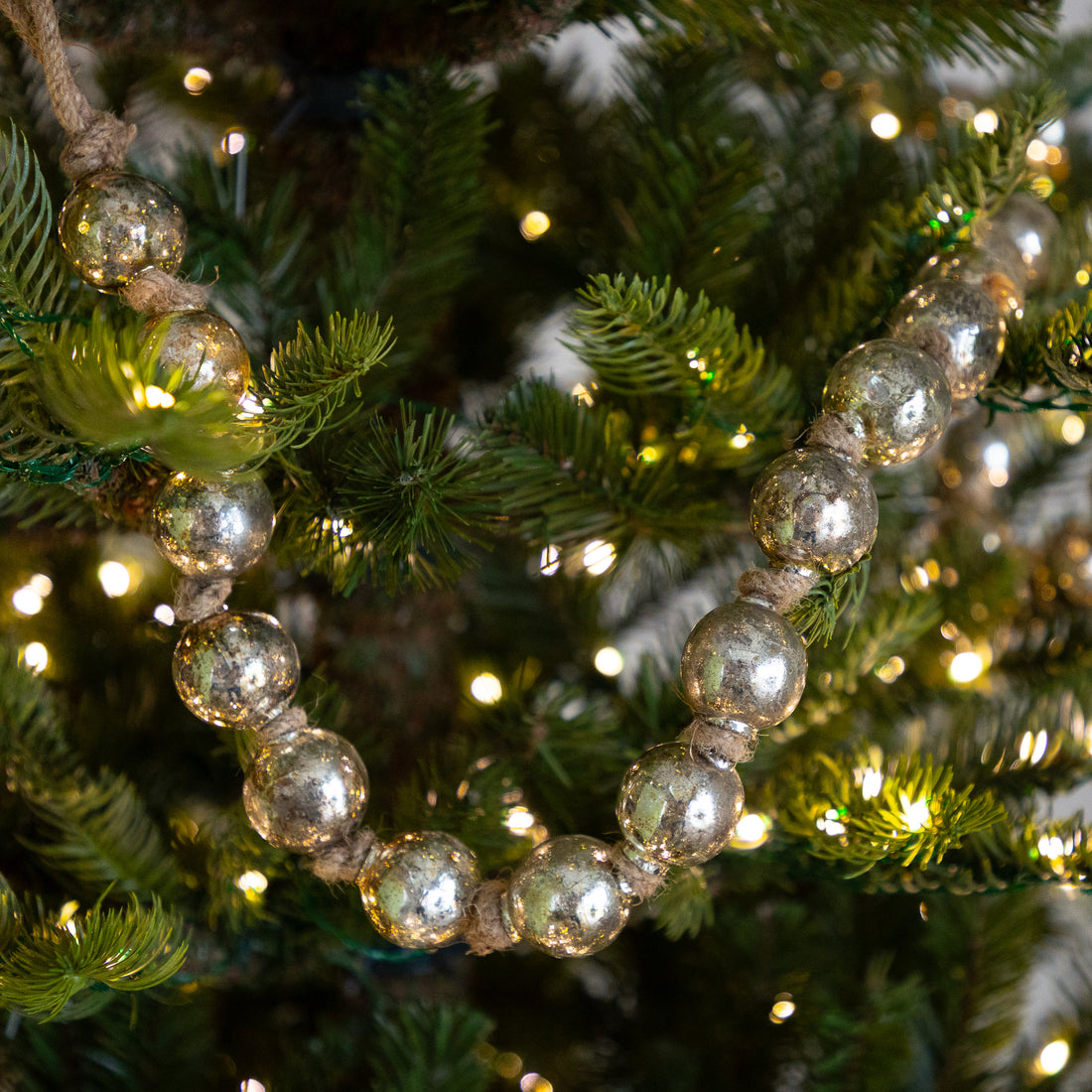 A strand of large, reflective silver glass beads wrapped on a rustic twine, draped over green pine tree branches with warm white string lights.