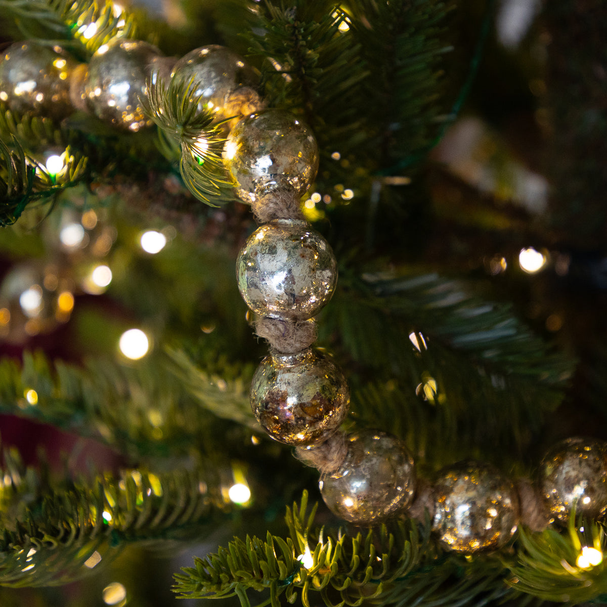 A close-up of a strand of metallic silver glass Christmas tree ornaments draped over green pine tree branches with warm white string lights glowing in the background.