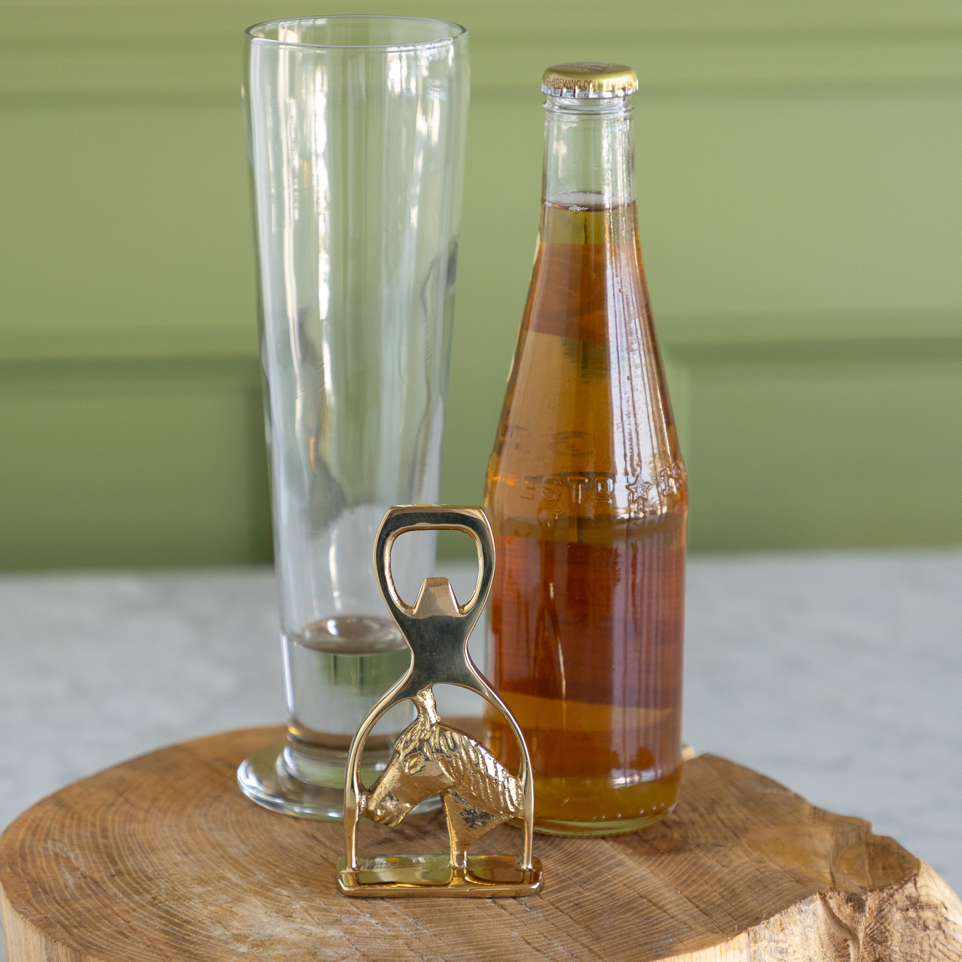 A brass bottle opener shaped like a horse stands on a round wooden slice in front of a clear glass and a glass soda bottle filled with amber liquid, set against a green paneled background.