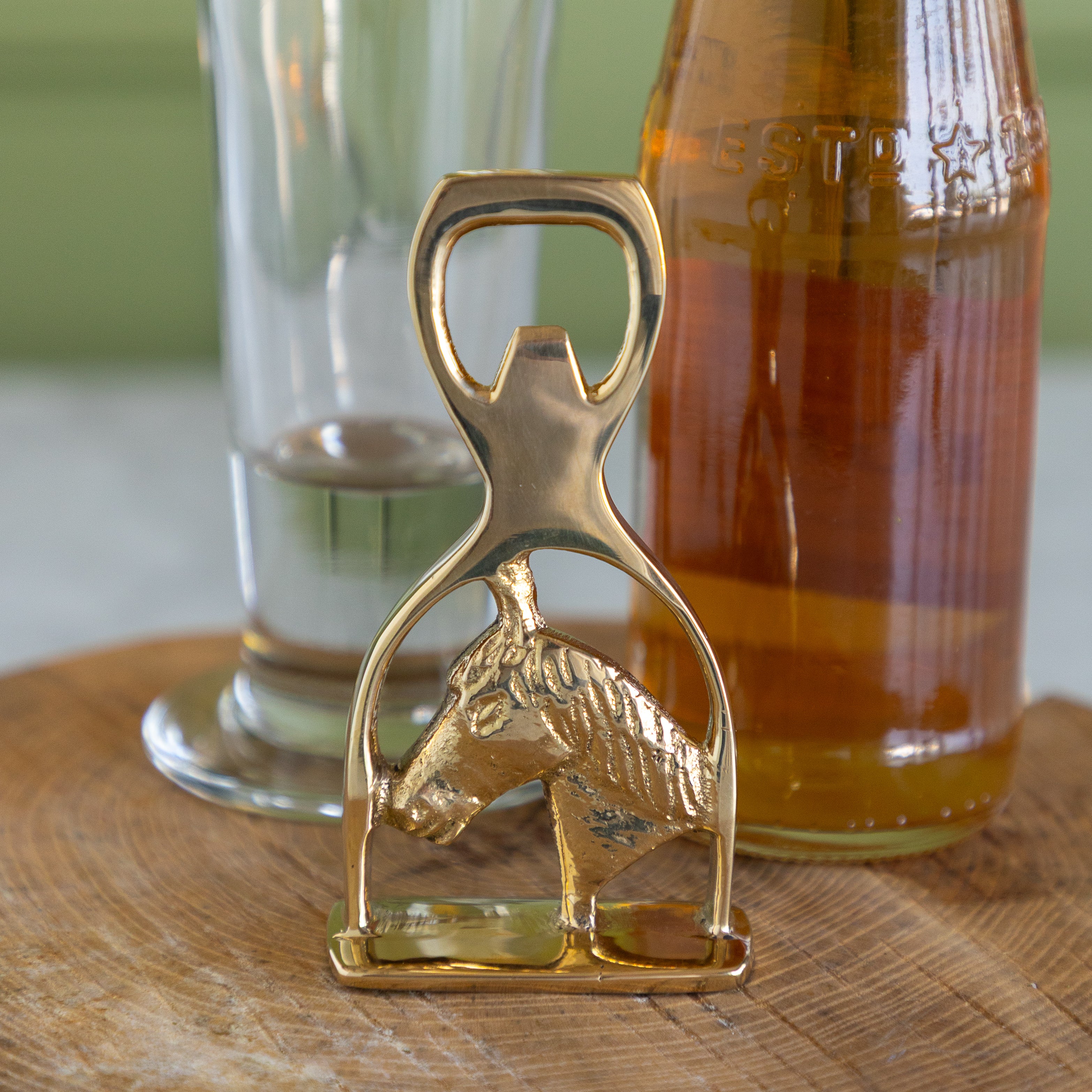 A gold-finished metal bottle opener shaped like a horse head, standing on a wooden surface with a glass and an amber bottle in the background.