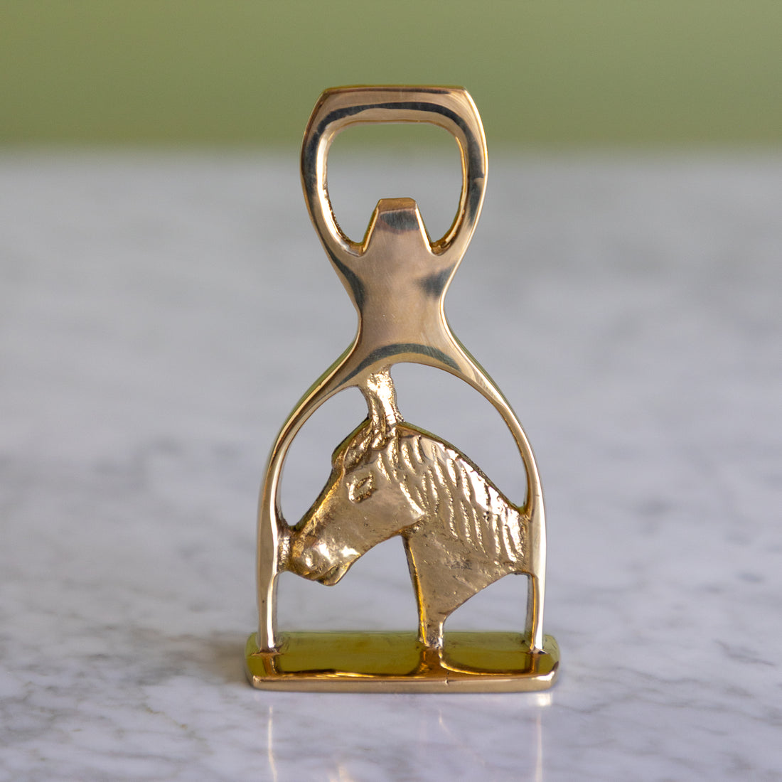 A polished gold-tone metal bottle opener shaped like a horse bit, featuring a textured horse head profile, resting on a light marble surface with a soft-focus green background.