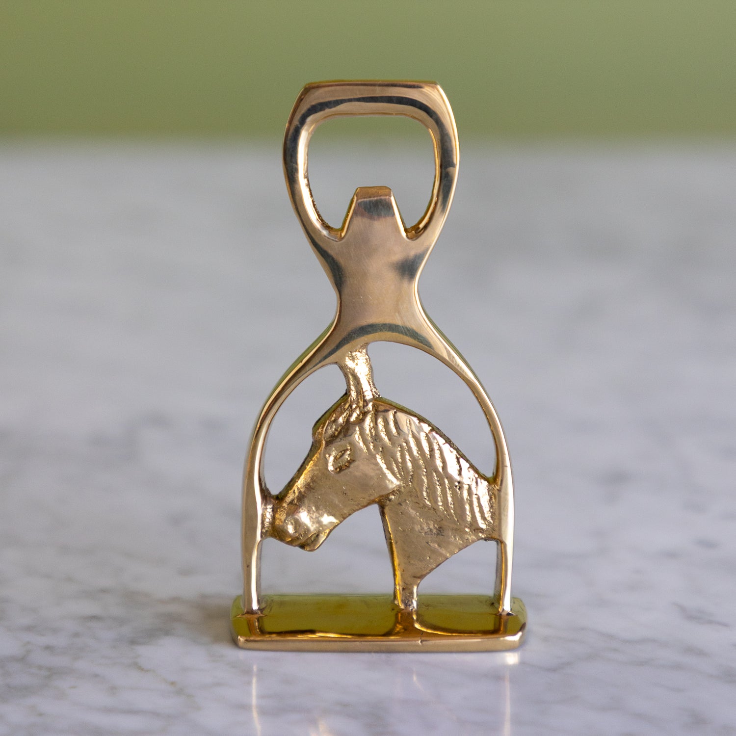 A polished gold-tone metal bottle opener shaped like a horse bit, featuring a textured horse head profile, resting on a light marble surface with a soft-focus green background.