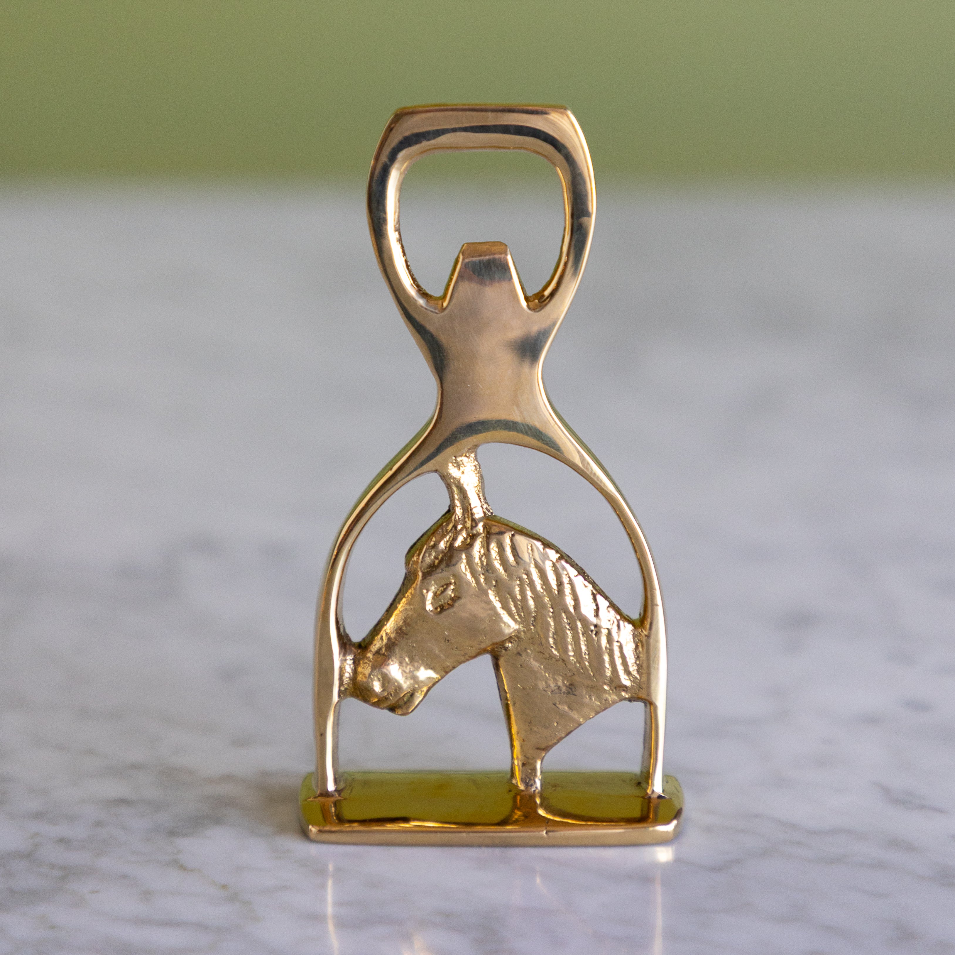 A polished gold-tone metal bottle opener shaped like a horse bit, featuring a textured horse head profile, resting on a light marble surface with a soft-focus green background.