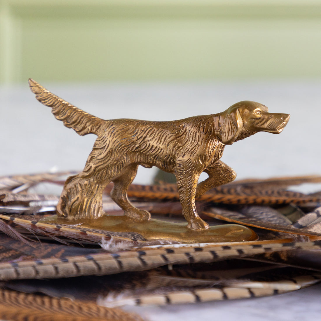 A small metallic dog figurine in a warm gold or brass tone, standing on a base decorated with brown and tan feathers, photographed against a softly blurred pale green and white background.