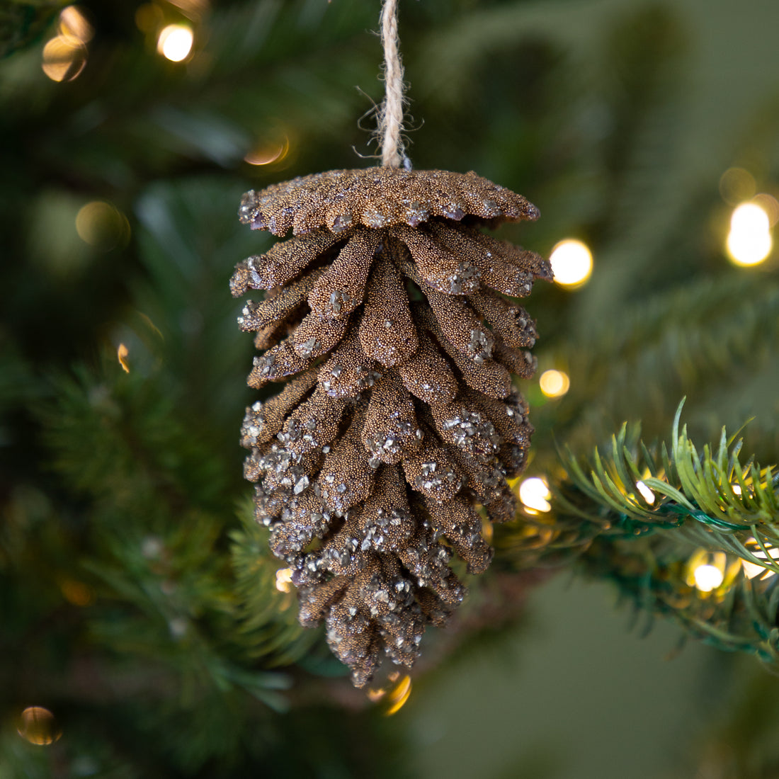 Decorative Beaded Glitter Pine Cone Ornament hanging on a Christmas tree with lights in the background.