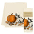 A cream-colored tea towel featuring an illustration of a black-and-white kitten next to an orange pumpkin with scattered autumn leaves, shown with its paper band packaging labeled Eric & CHRISTOPHER.