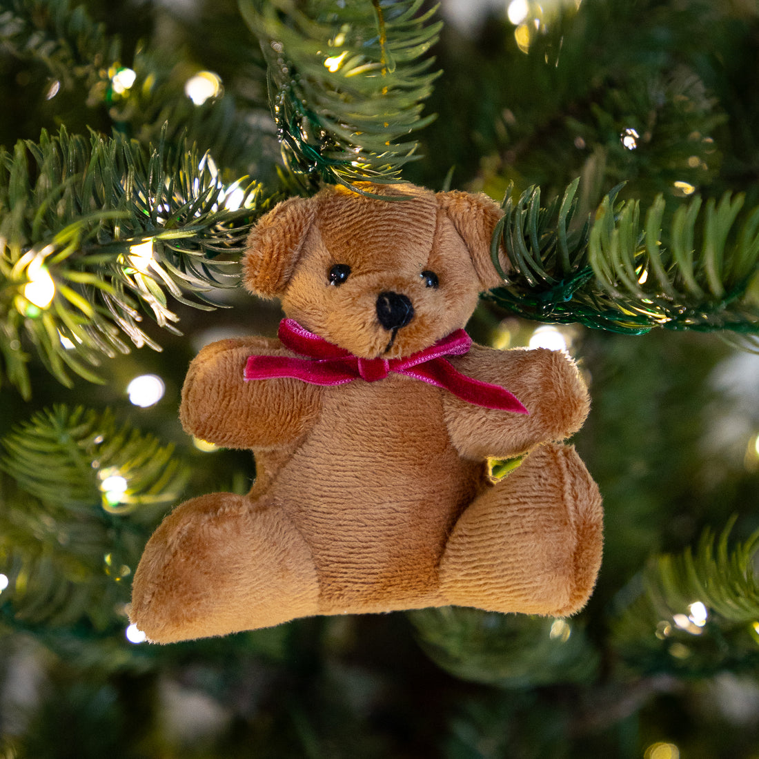 A brown plush teddy bear Christmas ornament with a pink ribbon bow, hanging on a green Christmas tree with warm white lights.