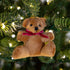 A brown plush teddy bear Christmas ornament with a pink ribbon bow, hanging on a green Christmas tree with warm white lights.
