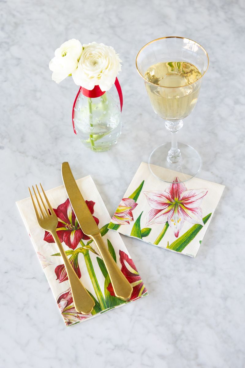 A square cocktail napkin and a rectangular guest napkin, both with red, pink and green florals, on a white marble tabletop with a glass of wine, a bud vase, and gold cutlery. 