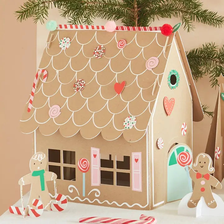 A gingerbread house craft decoration displayed on a tabletop with candy cane props and a Christmas tree branch in the background. The house is made from brown cardboard or paperboard with white icing-style lines, decorated with red and pink candy motifs, and small gingerbread figures stand beside it.