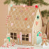 A gingerbread house craft decoration displayed on a tabletop with candy cane props and a Christmas tree branch in the background. The house is made from brown cardboard or paperboard with white icing-style lines, decorated with red and pink candy motifs, and small gingerbread figures stand beside it.