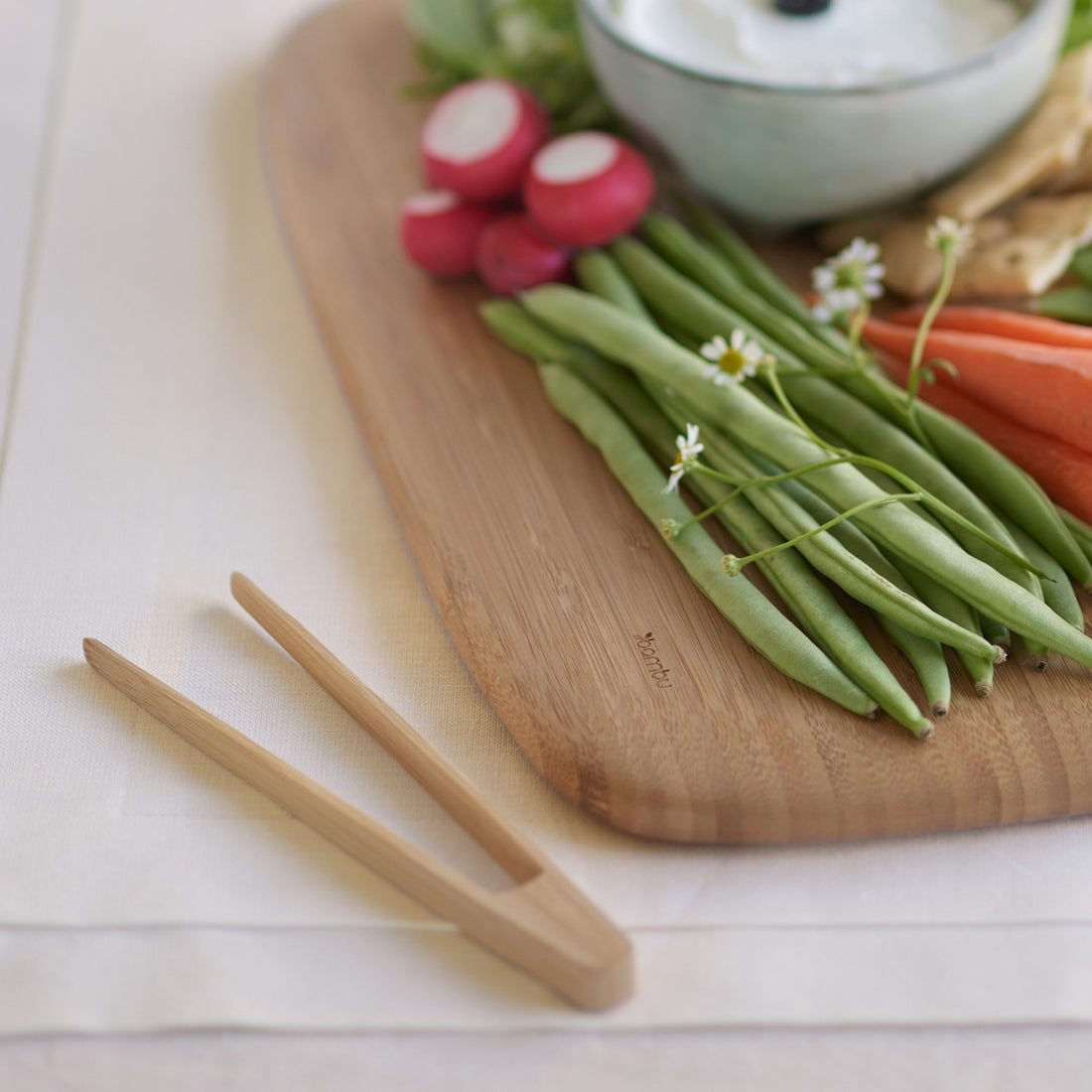 Wooden cutting board with green beans, carrots, radishes, and a bowl of dip on a light surface.