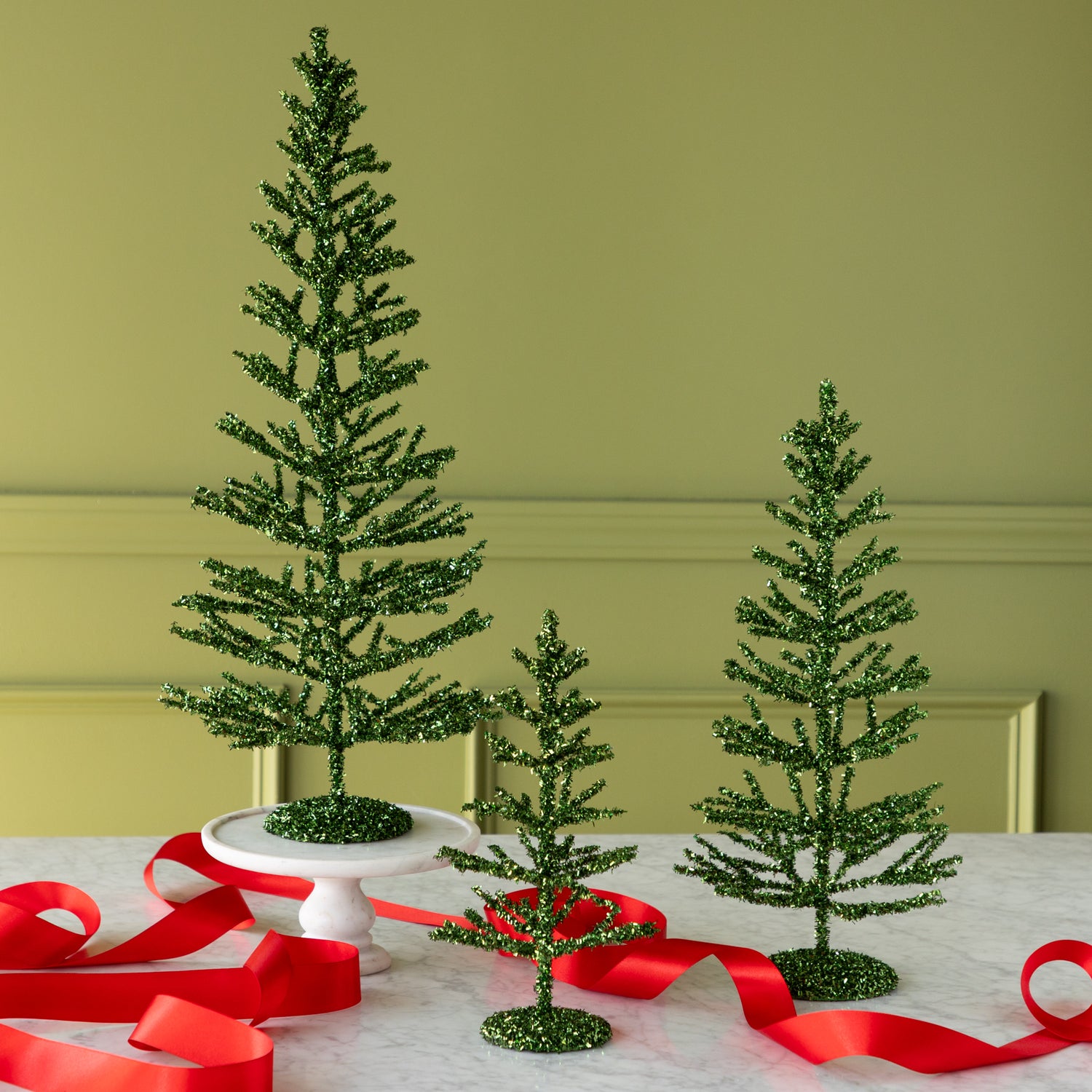 Three decorative green tinsel trees on a table with red ribbons against a green wall.