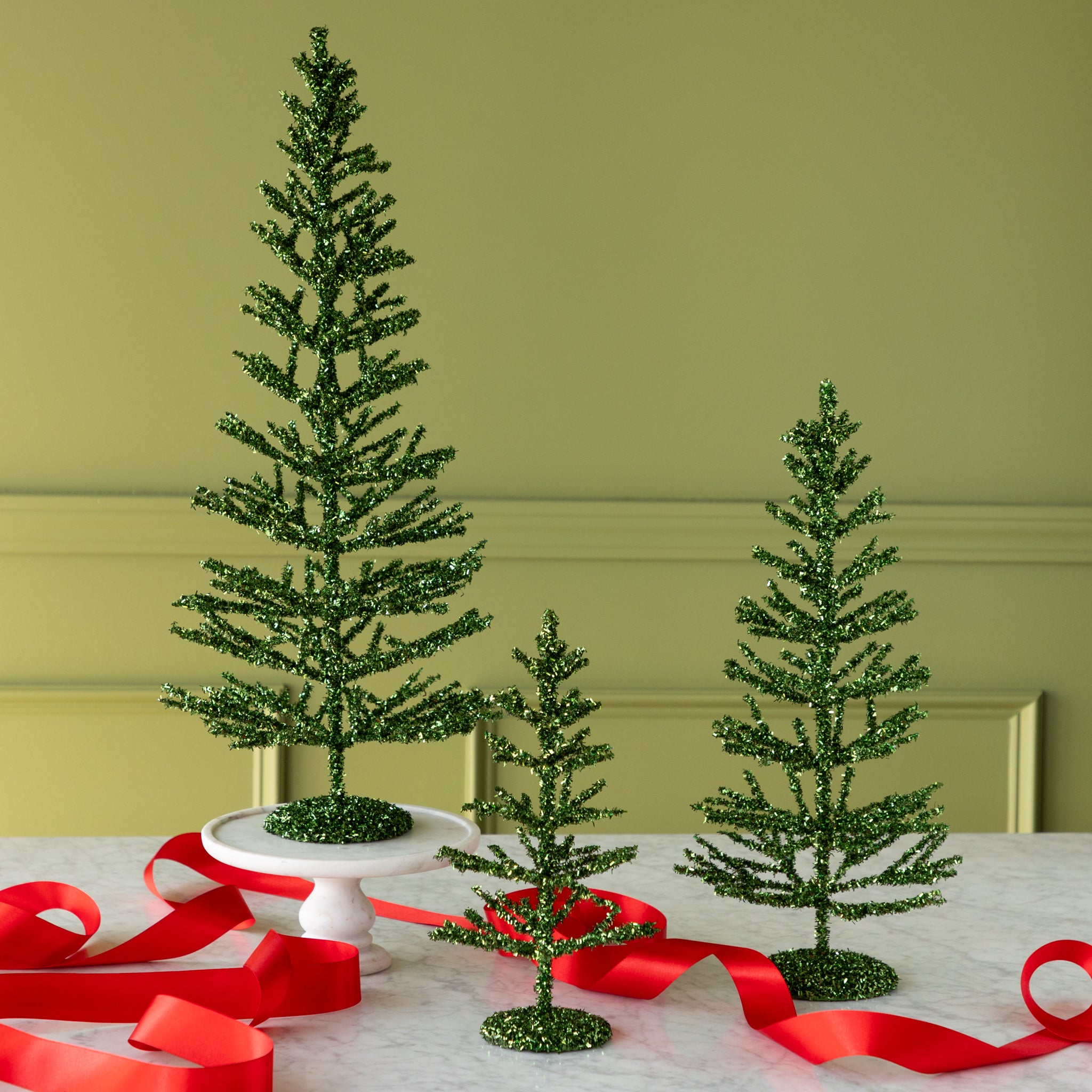 Three decorative green tinsel trees on a table with red ribbons against a green wall.