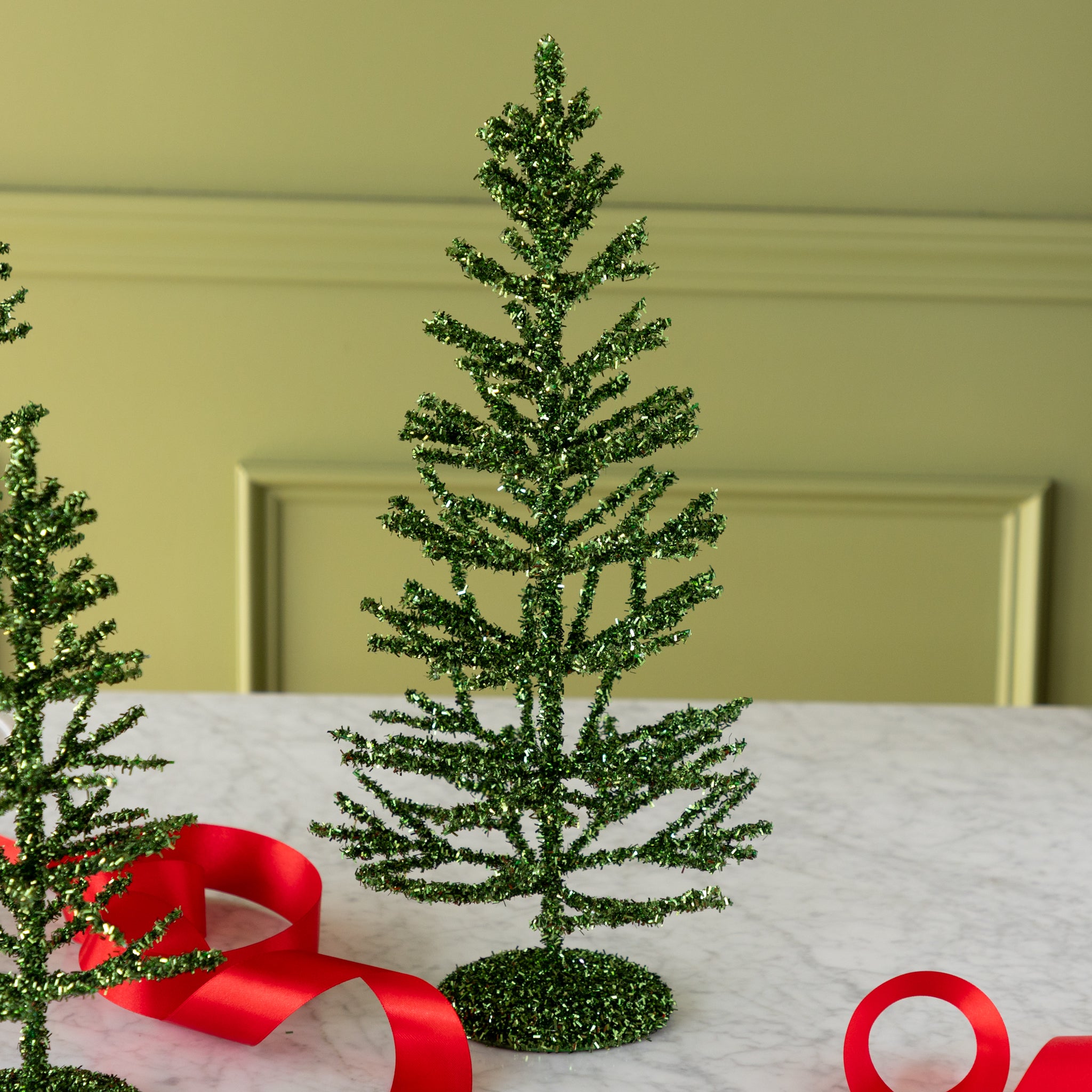 Small green artificial tinsel tree on a white surface with a red ribbon.