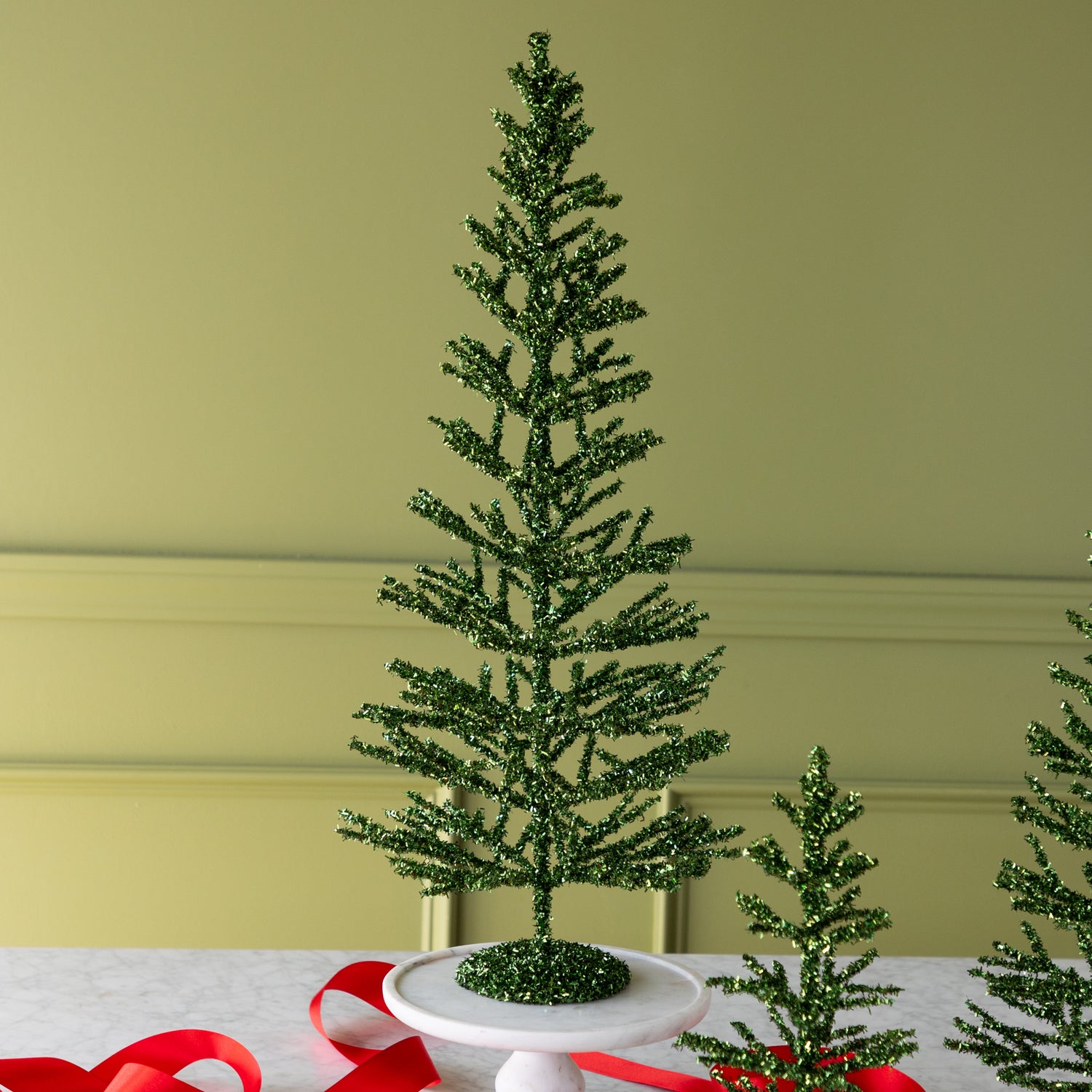 Large green artificial tinsel tree on a white surface with a red ribbon.