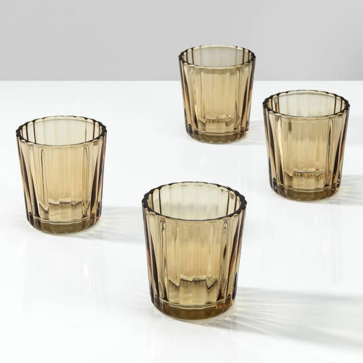 Four amber-tinted glass tumblers with a ribbed fluted design, arranged on a white reflective surface against a light gray background.
