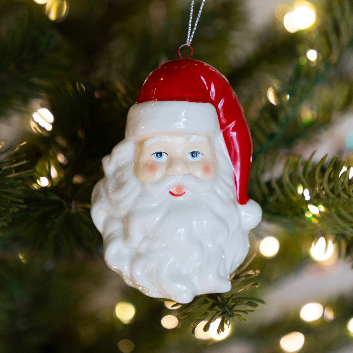 A glossy ceramic Santa Claus Christmas ornament hanging from a green Christmas tree branch with warm white bokeh lights in the background.
