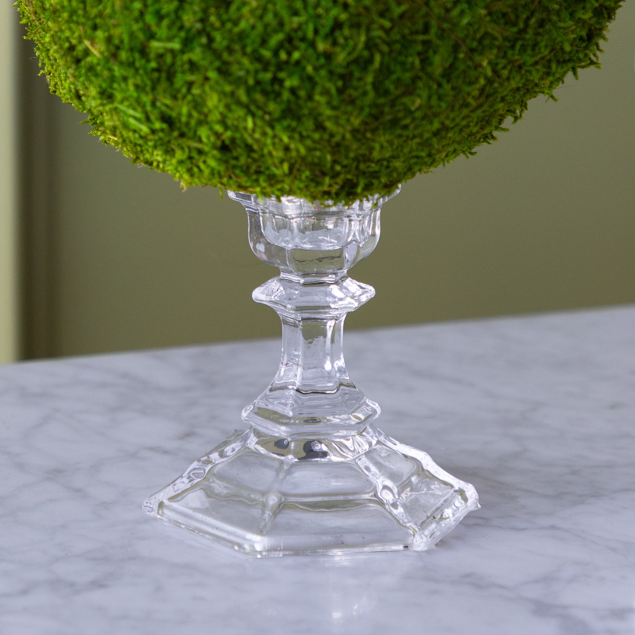 Decorative piece with green moss on a clear glass stand on a marble surface.