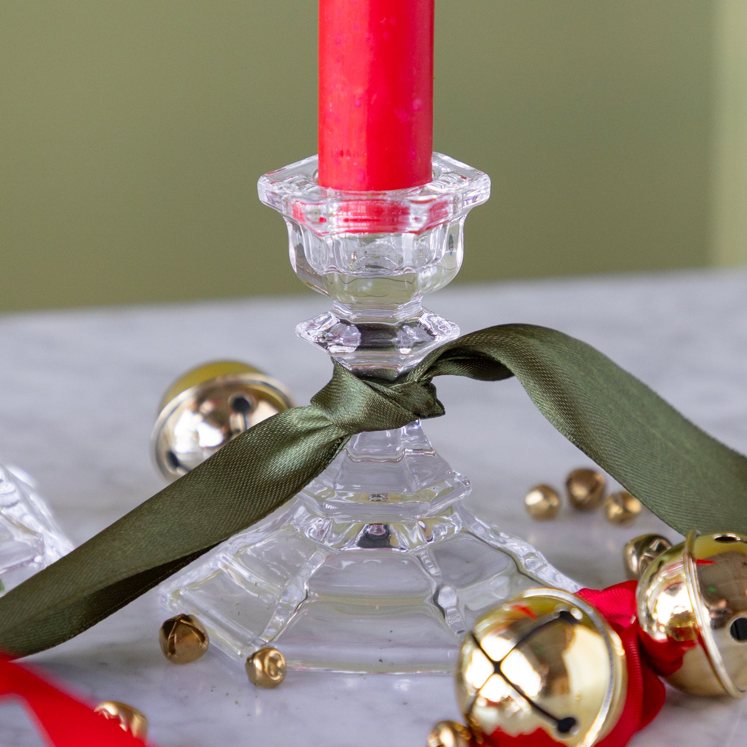 Clear glass candle holder with a red candle, green ribbon, and gold bells on a white surface.