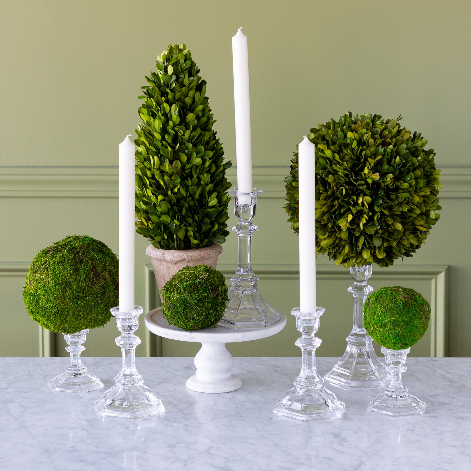Decorative arrangement with green spherical topiaries and white candles on a marble surface.