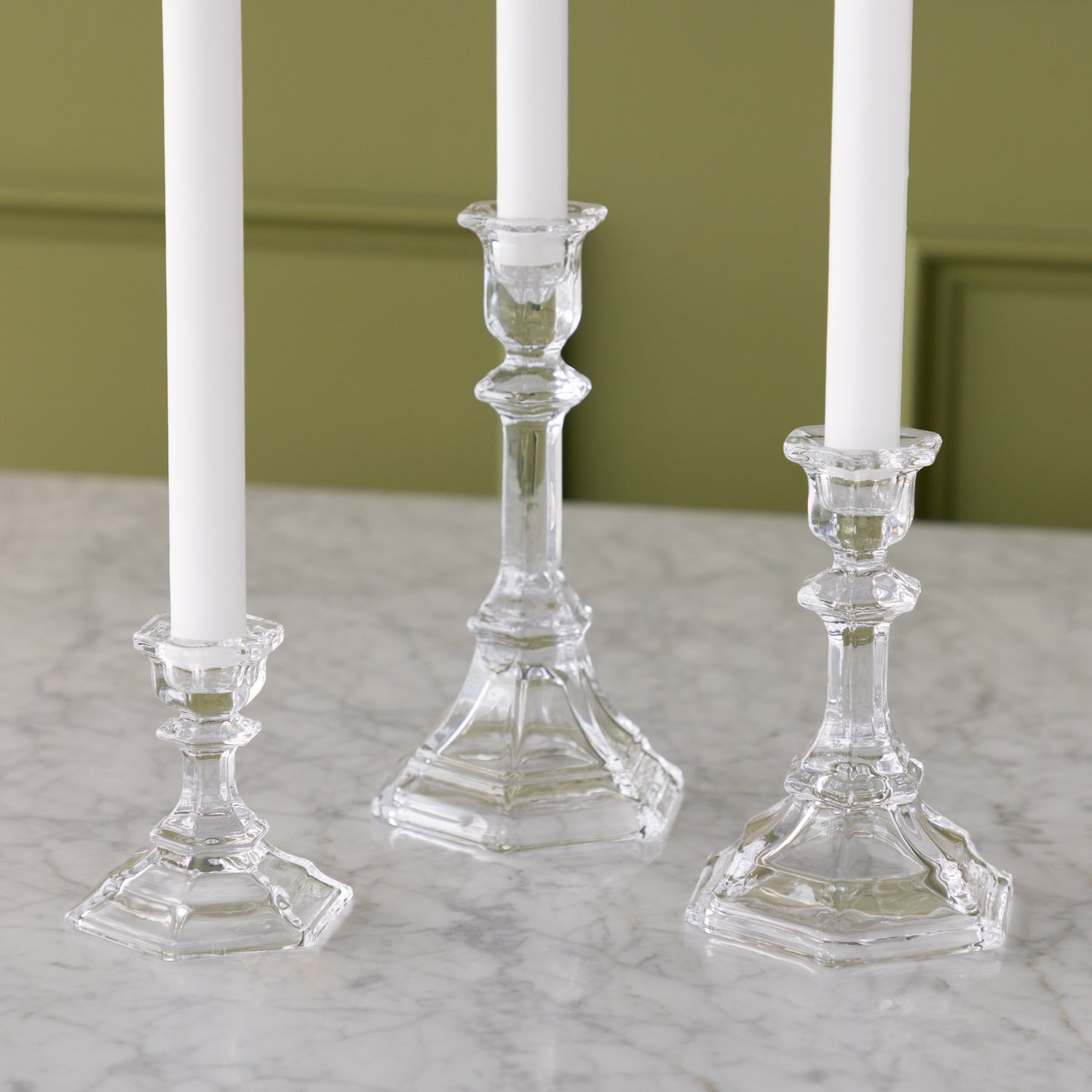 Three clear glass candlesticks with white candles on a marble surface against a green wall.