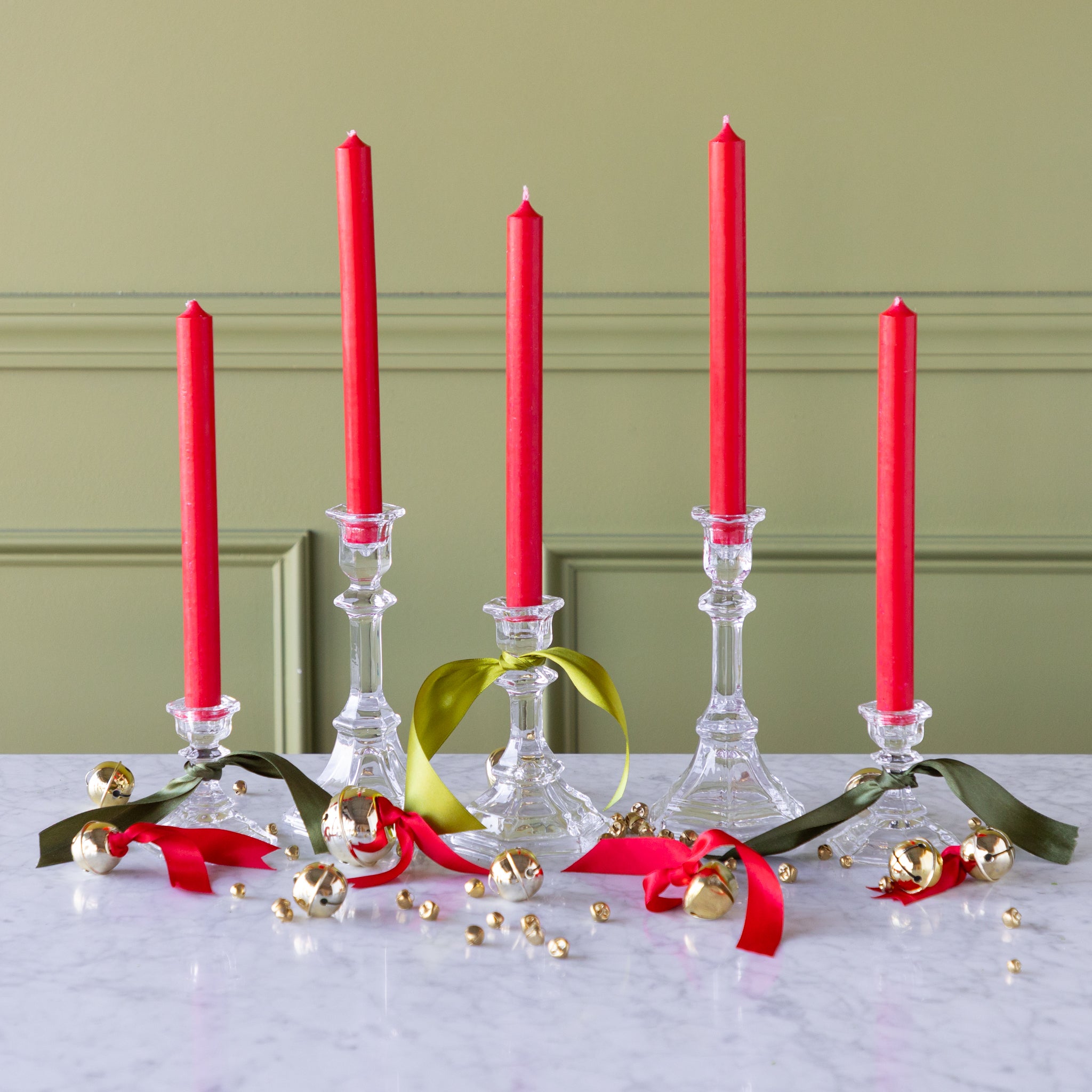 Red candles in glass candlesticks with decorative ribbons and bells on a marble surface against a green wall.