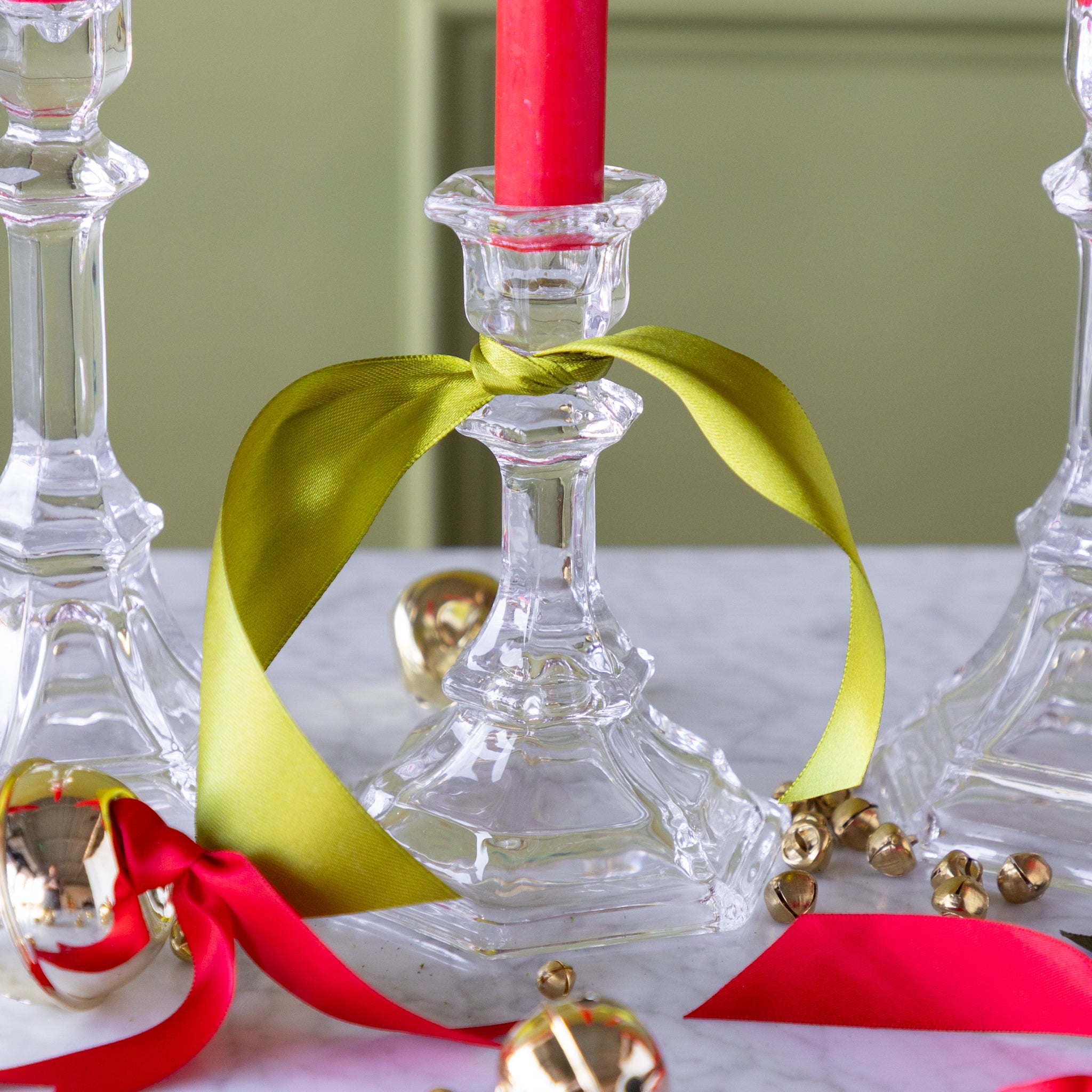 Clear glass candlestick with a red candle and green ribbon on a light background.