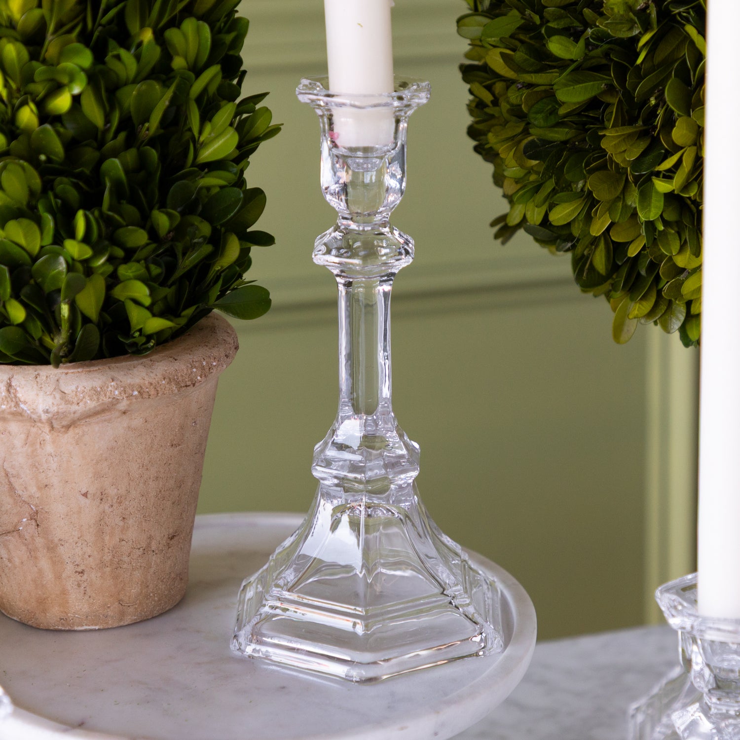 Clear glass candlestick with a white candle, potted plant, and greenery on a marble surface.