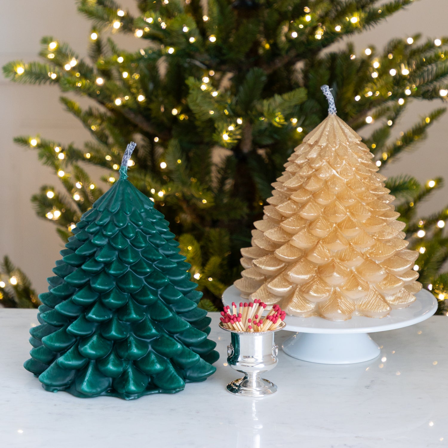 Two Christmas tree-shaped candles, one green and one gold, on a table next to a container of matches with a Christmas tree in the background.
