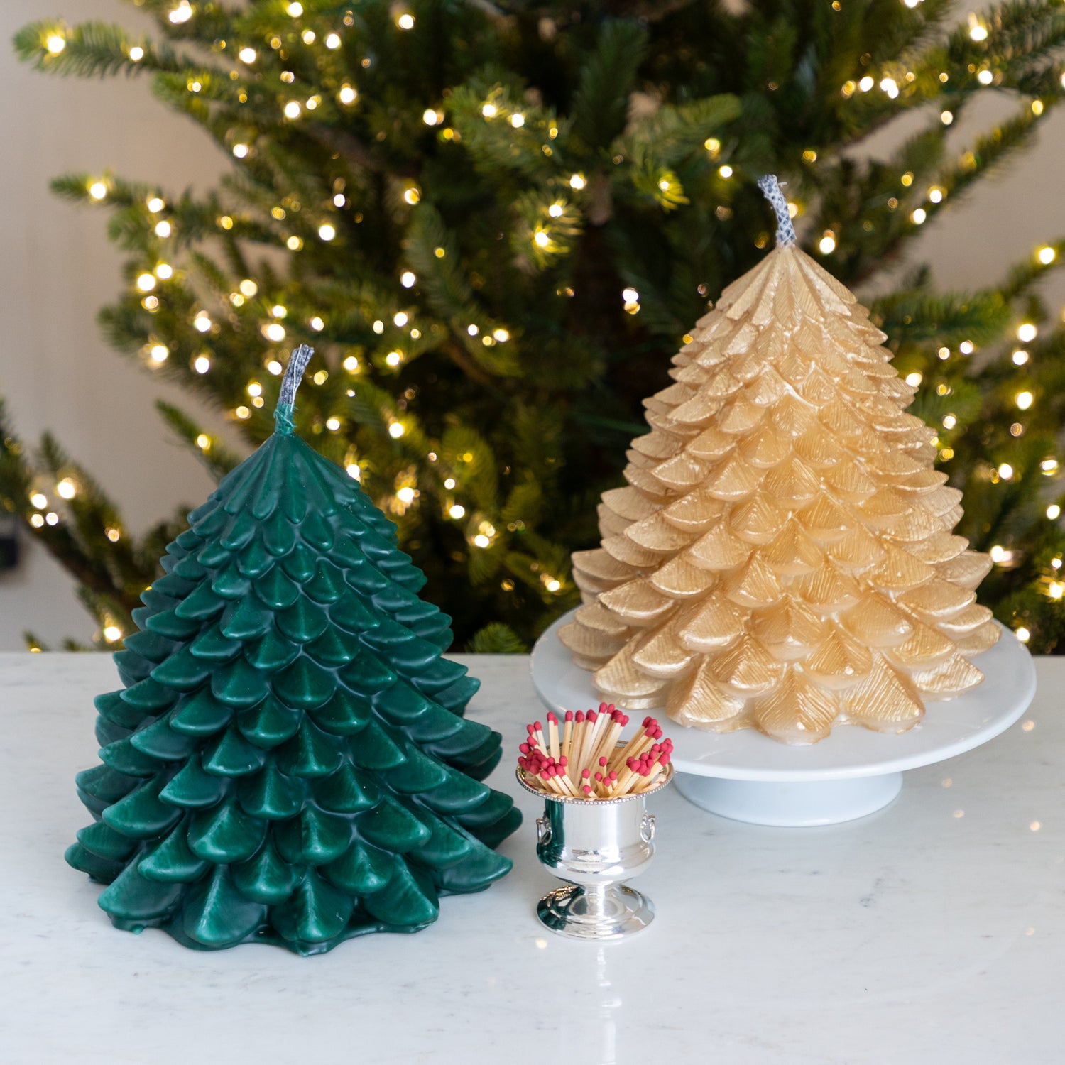 Two Christmas tree-shaped candles, one green and one gold, on a table next to a container of matches with a Christmas tree in the background.