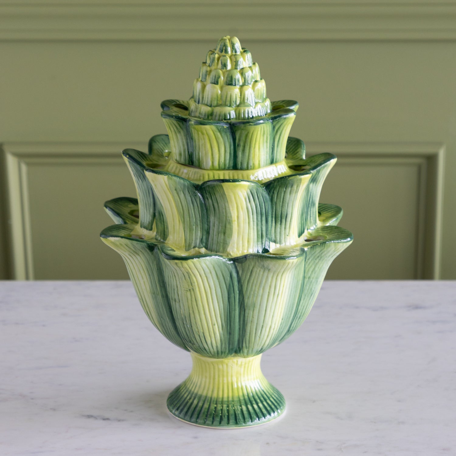 Green ceramic vase with tiered design on a white surface against a green wall.