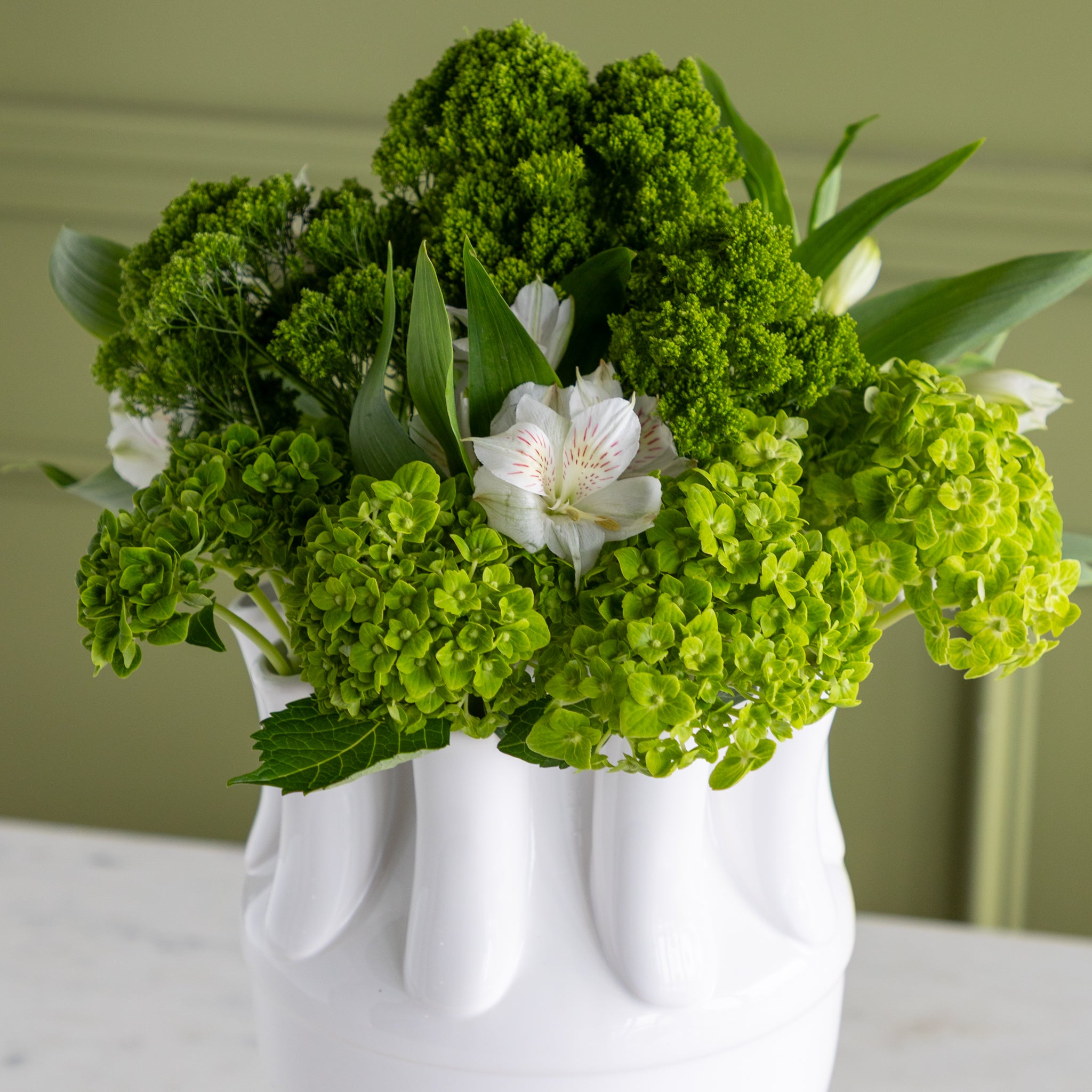 Bouquet of green and white flowers in a white vase on a light surface with a green background.