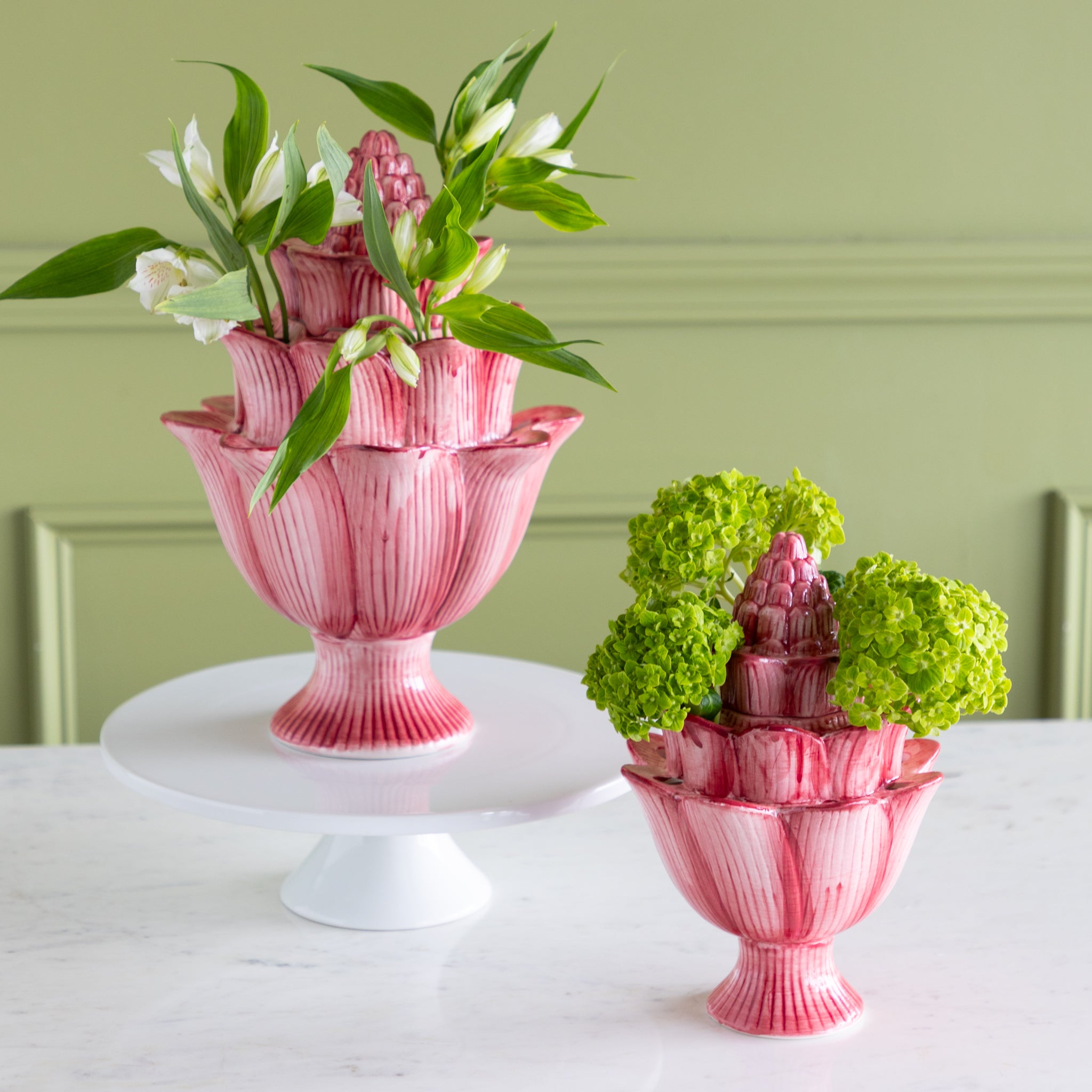 Pink decorative vases with floral arrangements on a white surface against a green wall.