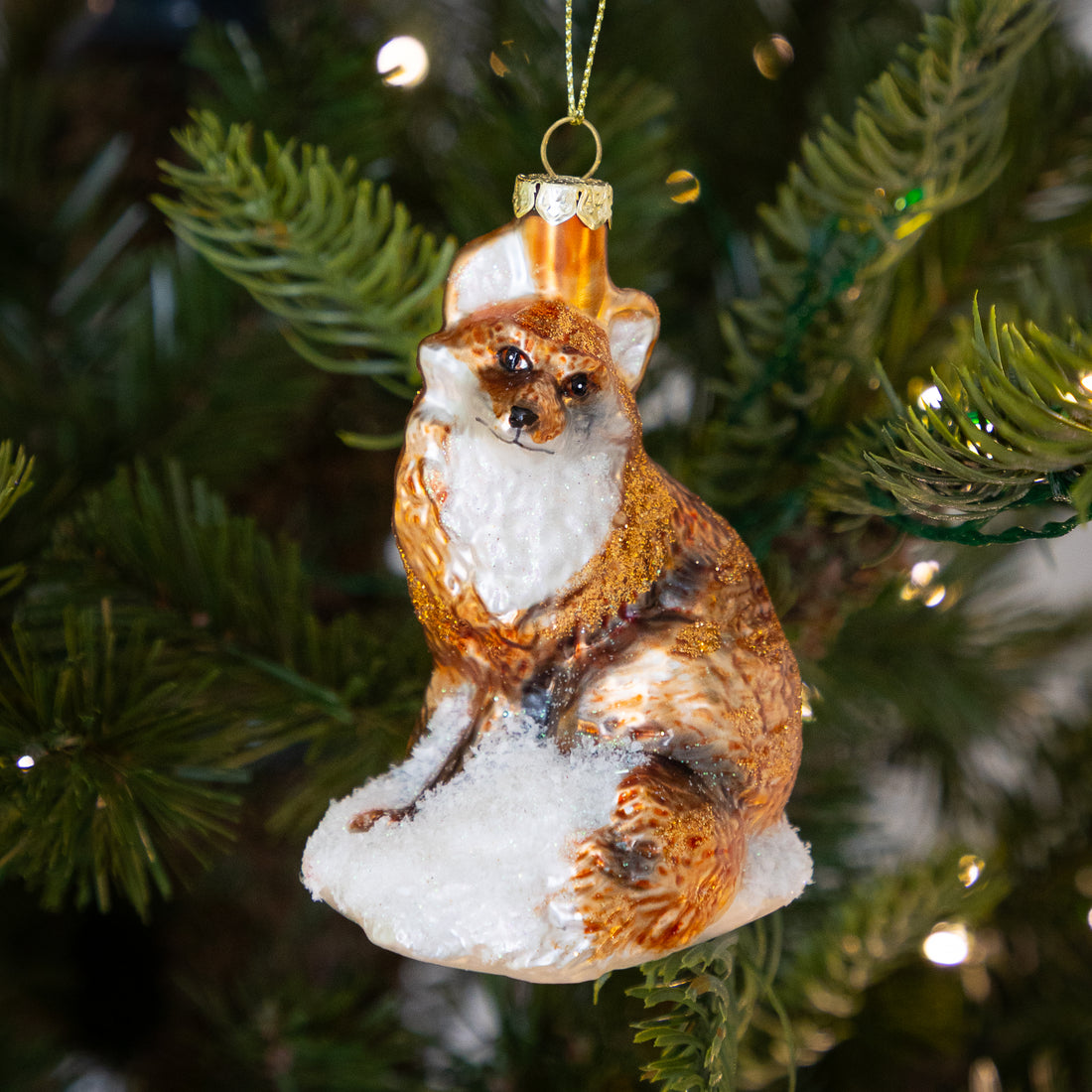 A hand-painted glass fox Christmas ornament hanging on a green Christmas tree branch with warm string lights in the background.