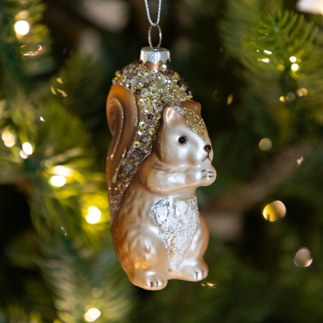 A glass squirrel Christmas ornament with a brown and silver body, glittery gold tail, and a silver heart on its chest, hanging in front of a blurred green Christmas tree with warm lights.