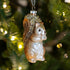 A glass squirrel Christmas ornament with a brown and silver body, glittery gold tail, and a silver heart on its chest, hanging in front of a blurred green Christmas tree with warm lights.