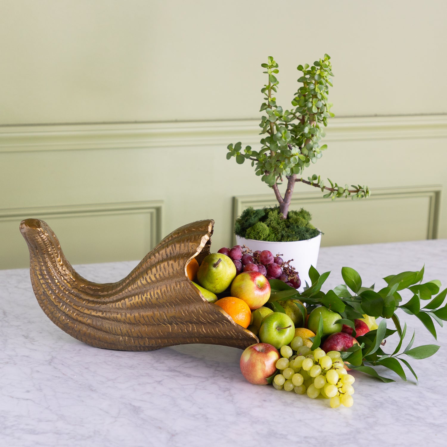 A decorative gold cornucopia-style bowl filled with fruit, placed on a white marble table in front of a potted green plant against a pale green paneled wall.