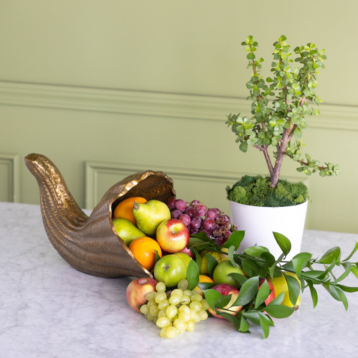 A decorative gold-toned cornucopia-style fruit bowl filled with oranges, green apples, red apples, and purple grapes, styled on a marble surface beside a potted green tree against a pale green paneled wall.