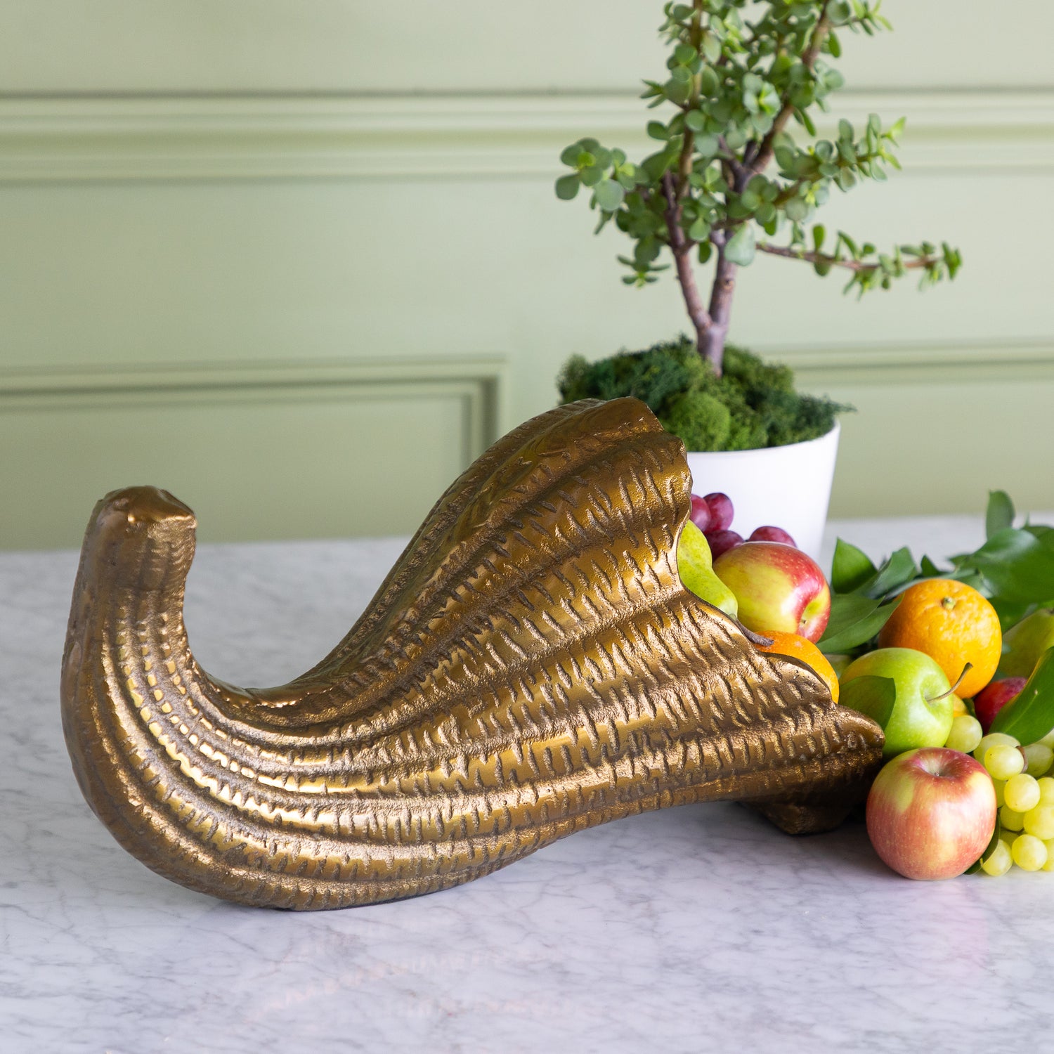 A decorative gold cornucopia-shaped fruit bowl filled with apples, oranges, and grapes, styled on a marble surface with a potted plant and paneled wall in the background.
